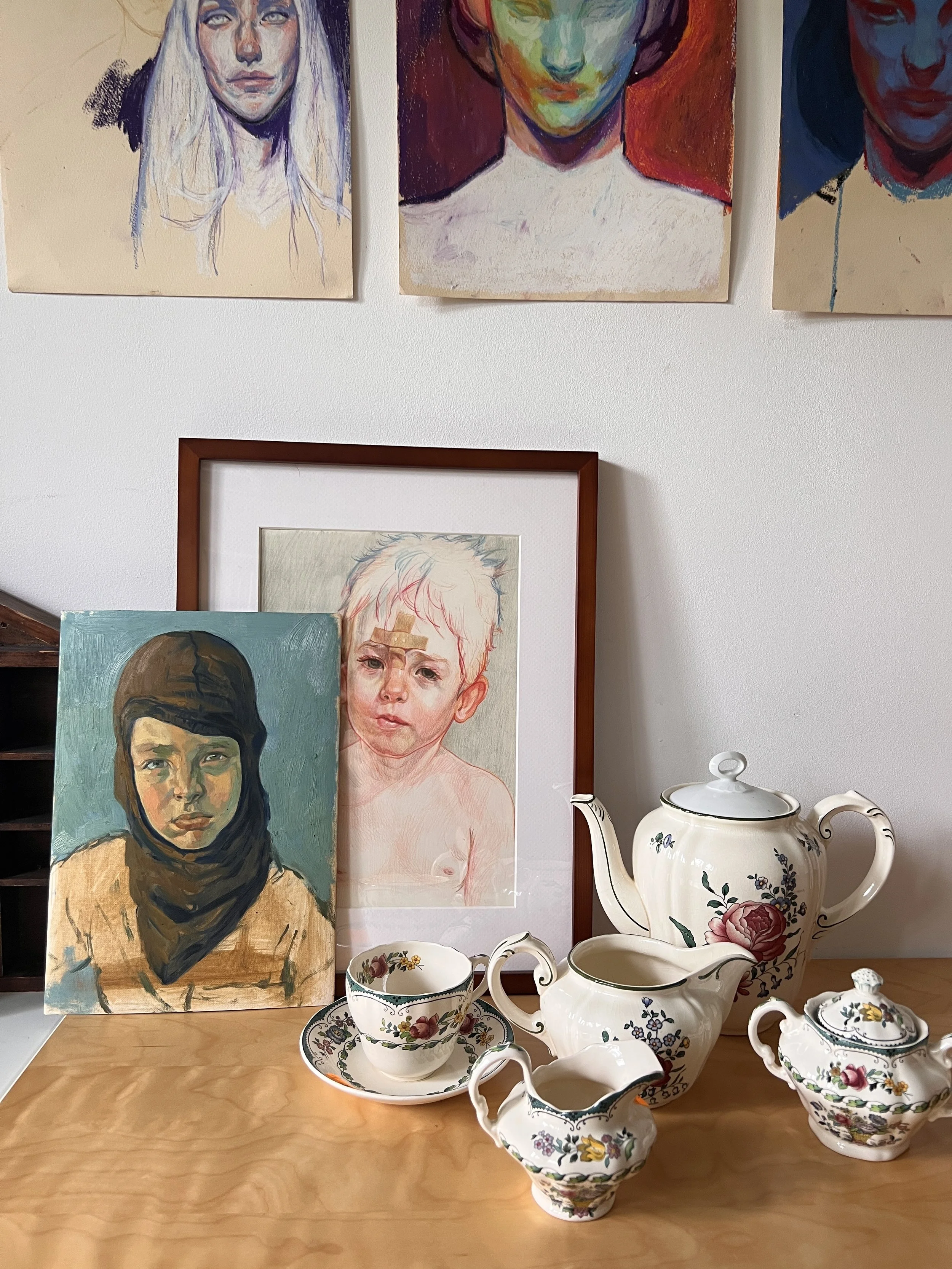 A still life scene with decorative teapots and cups on a wooden surface, framed artwork of two children, and three portrait paintings hanging on the wall above.
