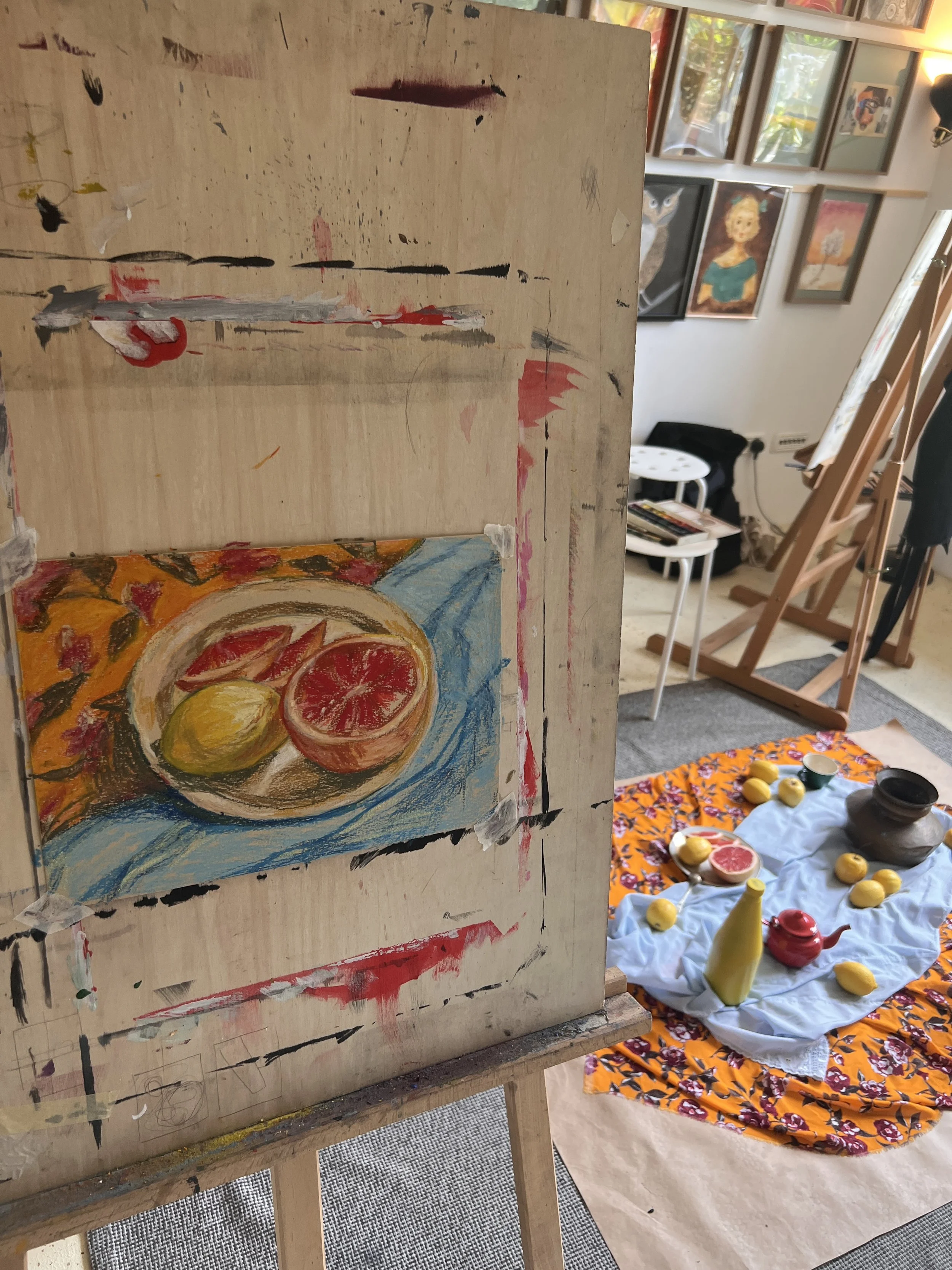 An artist's painting of a still life featuring oranges and lemons on a plate, with a background of orange and pink flowers and a blue cloth. The painting is on an easel in an art studio.