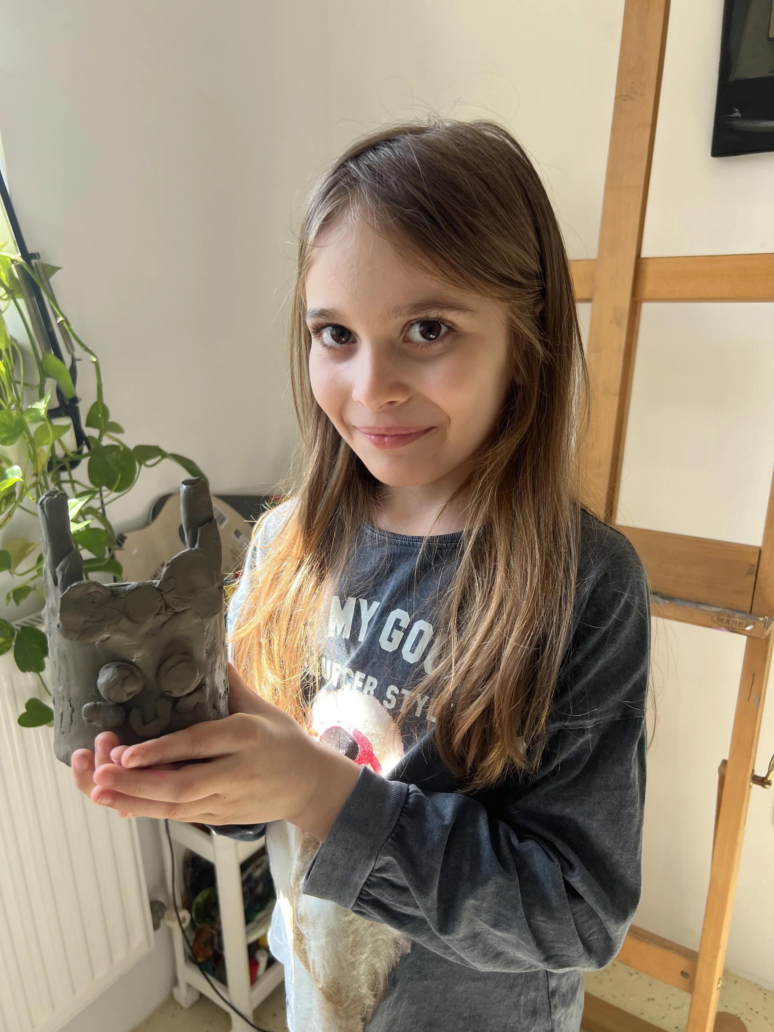 A young girl with long light brown hair, smiling, holding a gray ceramic sculpture of a face with horns.