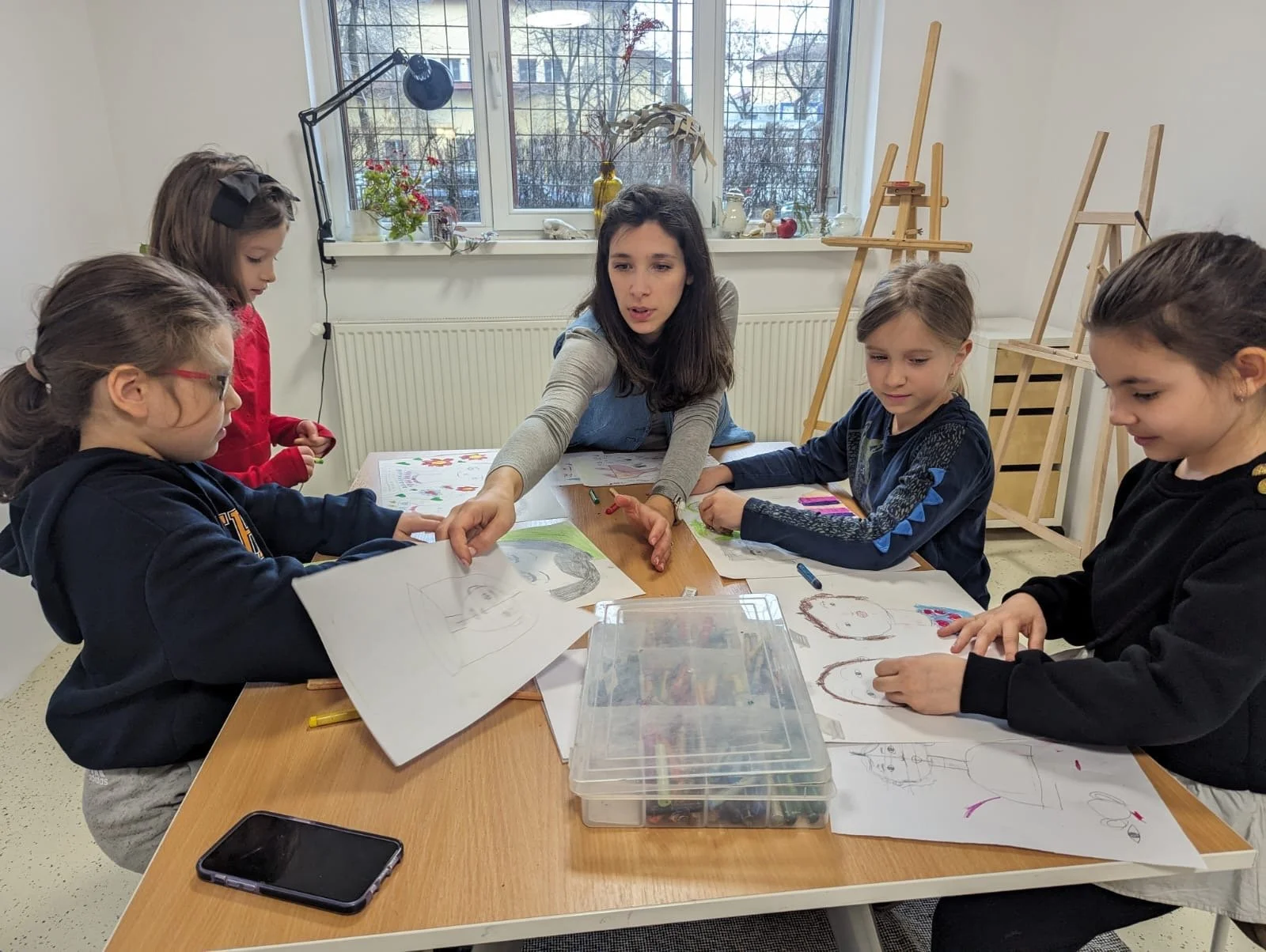 A teacher and four young girls are sitting around a table in an art classroom, working on drawings and art projects.