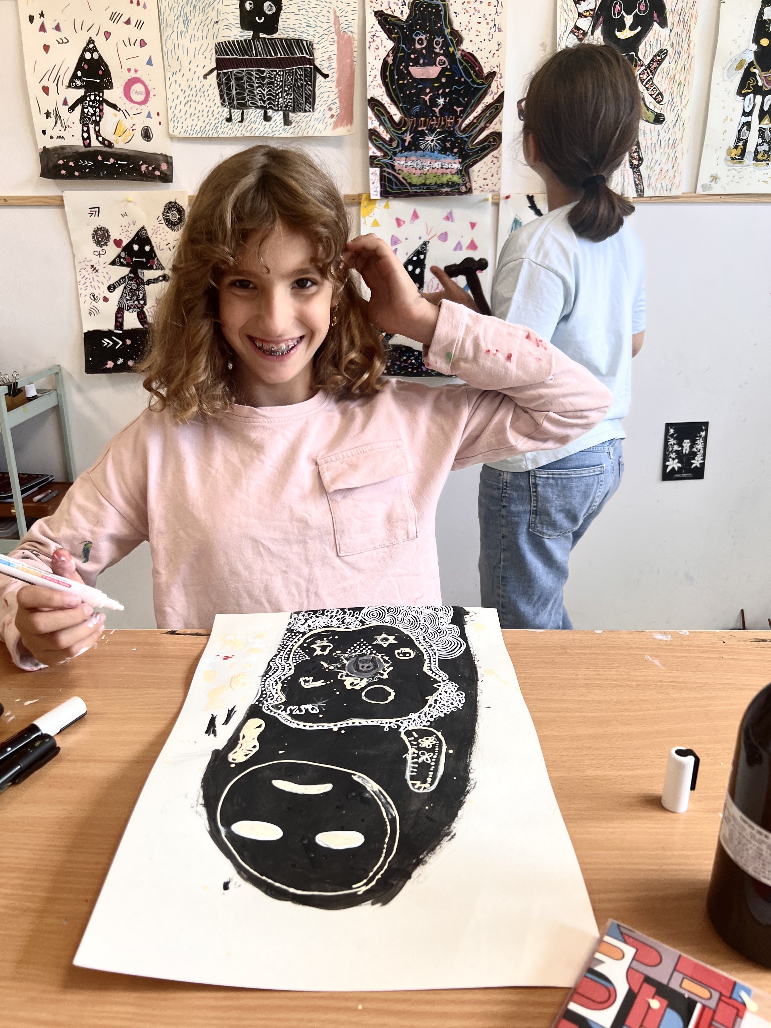 A young girl with curly hair and braces smiling at the camera while sitting at a table with her artwork, a black and white print of a face. Behind her on the wall are various colorful paintings of animals and abstract designs. Another girl with straight hair tied back, wearing a light blue shirt, is standing and working on her art.