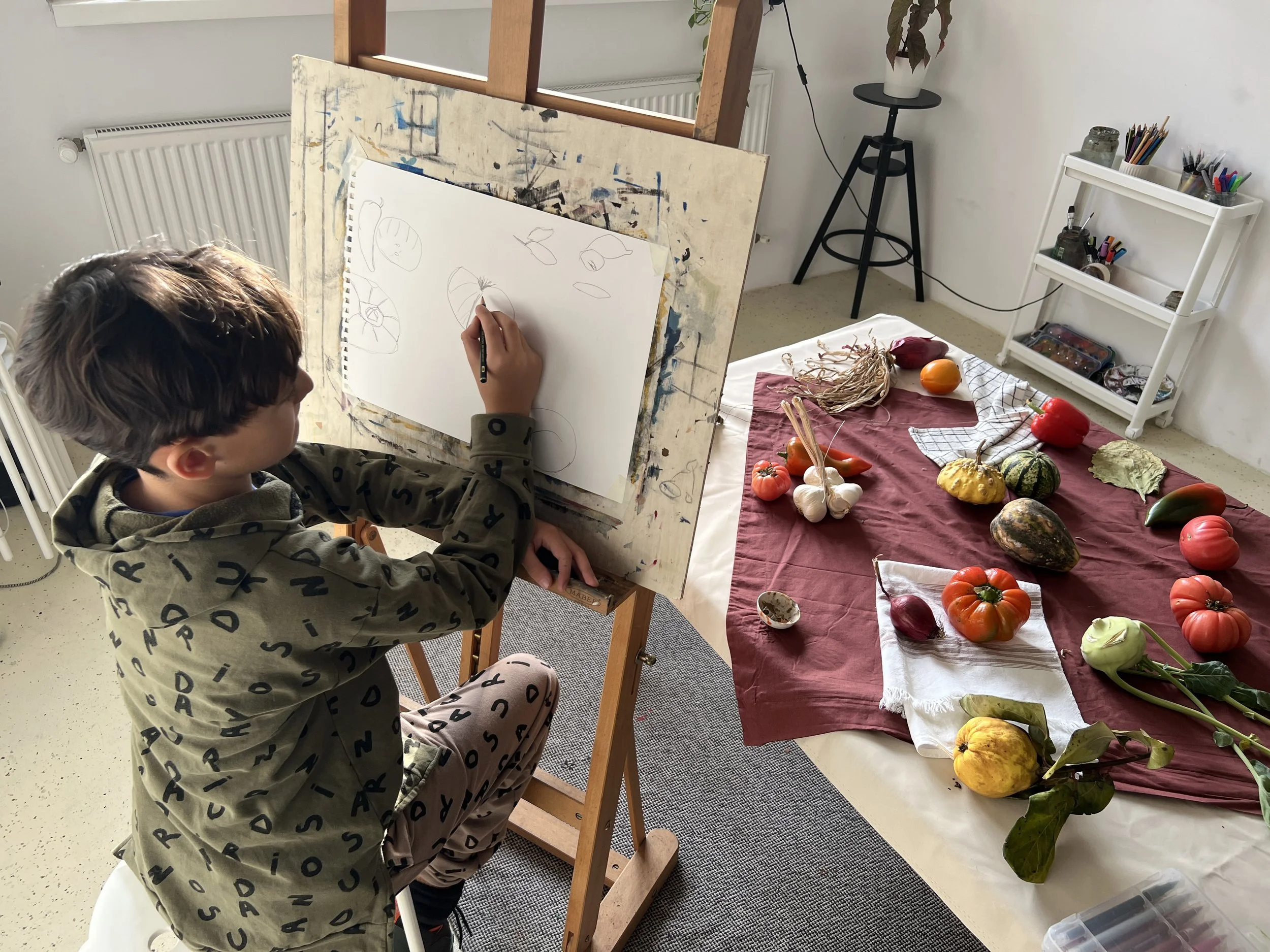 A child sitting on a stool drawing on a large sheet of paper mounted on an easel. The paper is blank except for a few simple sketches. In front of him, a table is covered with a red cloth and topped with various vegetables and gourds. A small white towel and some leaves are also on the table. In the background, there is a white shelf with art supplies and a small plant in a pot.