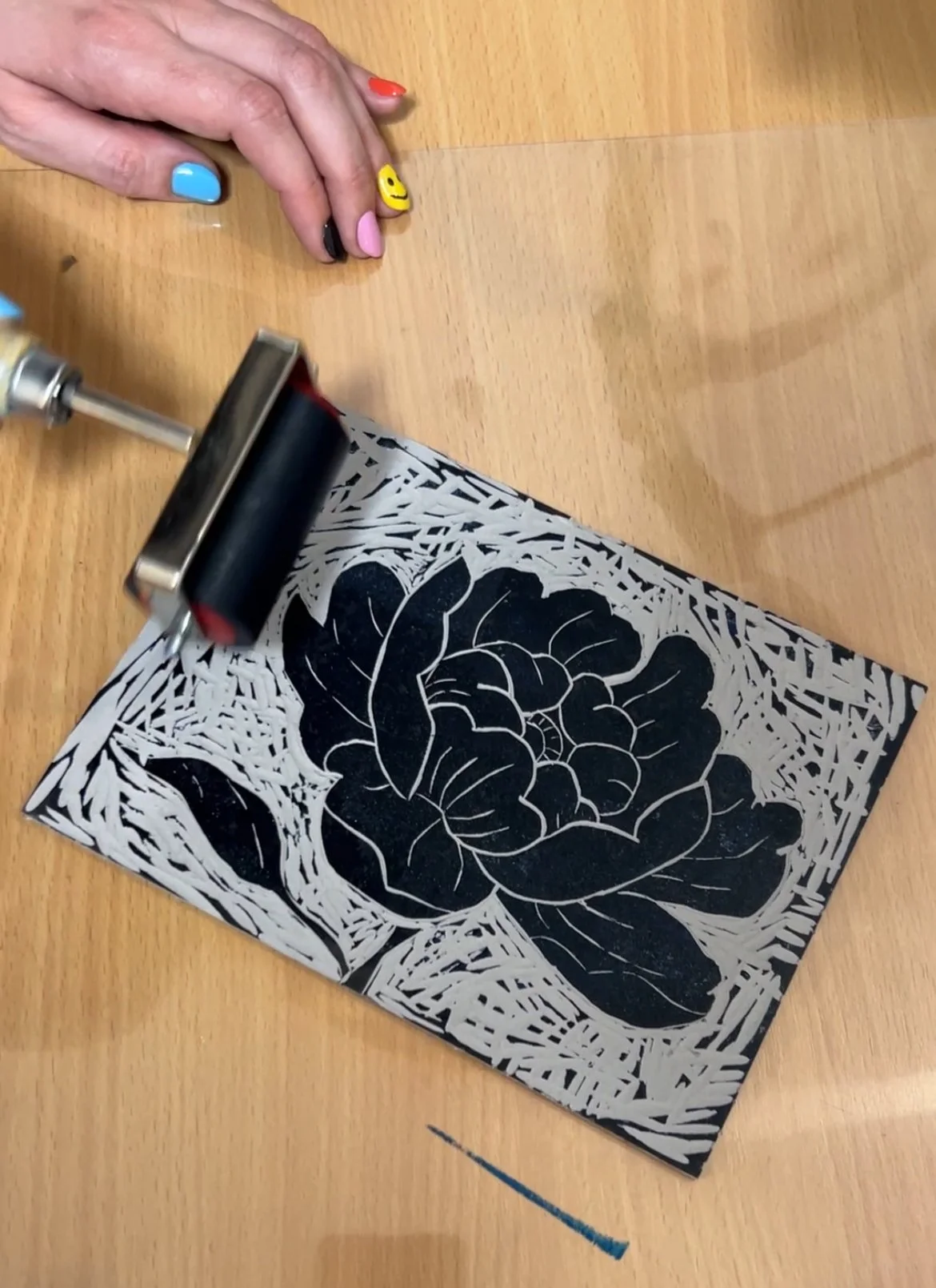 A person with colorful and decorated nails presses a roller against a black-and-white floral paper cutout, possibly for a printing or craft project, on a wooden surface.