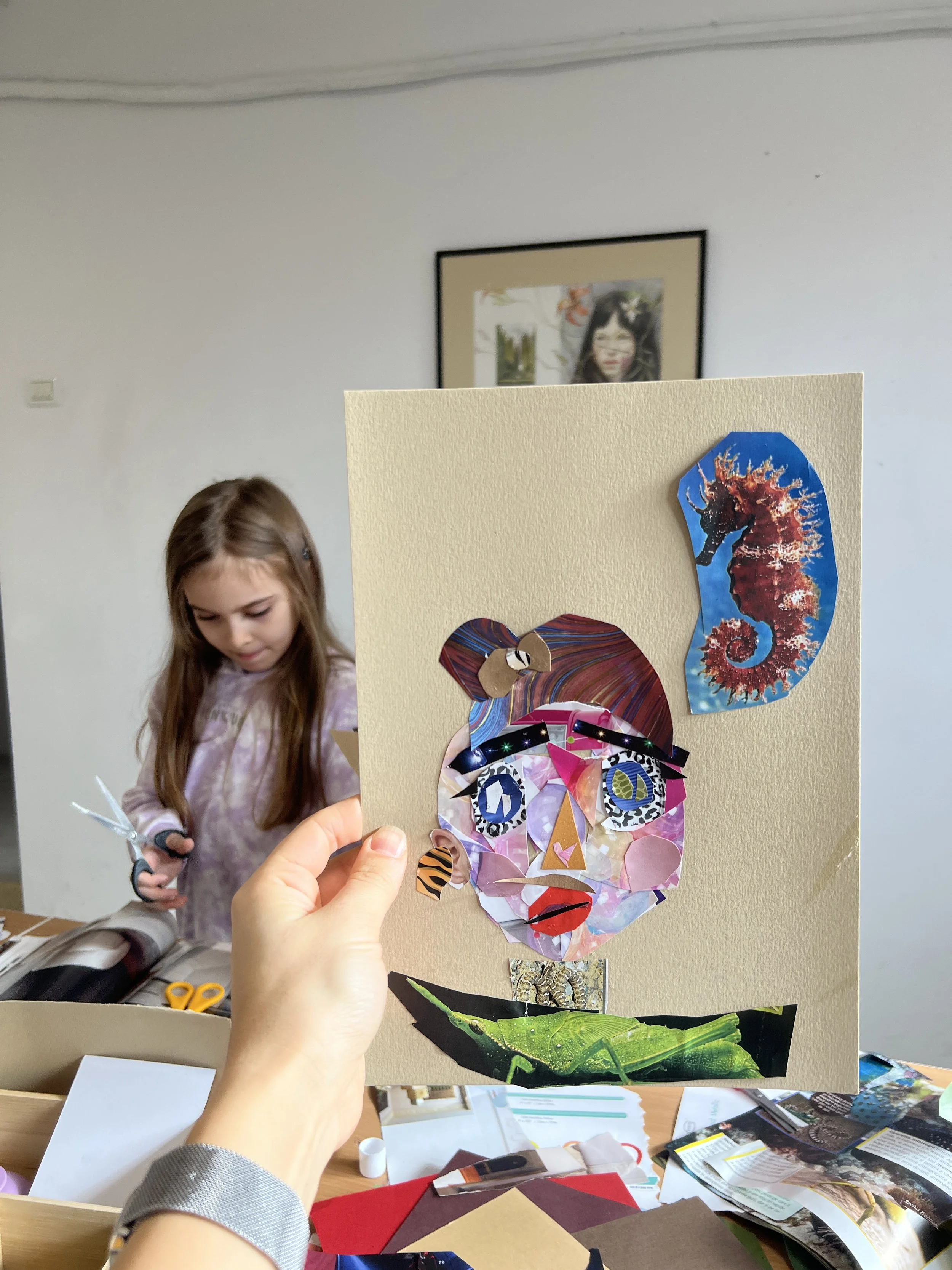 Hand holding up a collage art piece of a human face with an ocean and seahorse theme, surrounded by art supplies on a table and a girl in the background holding scissors.