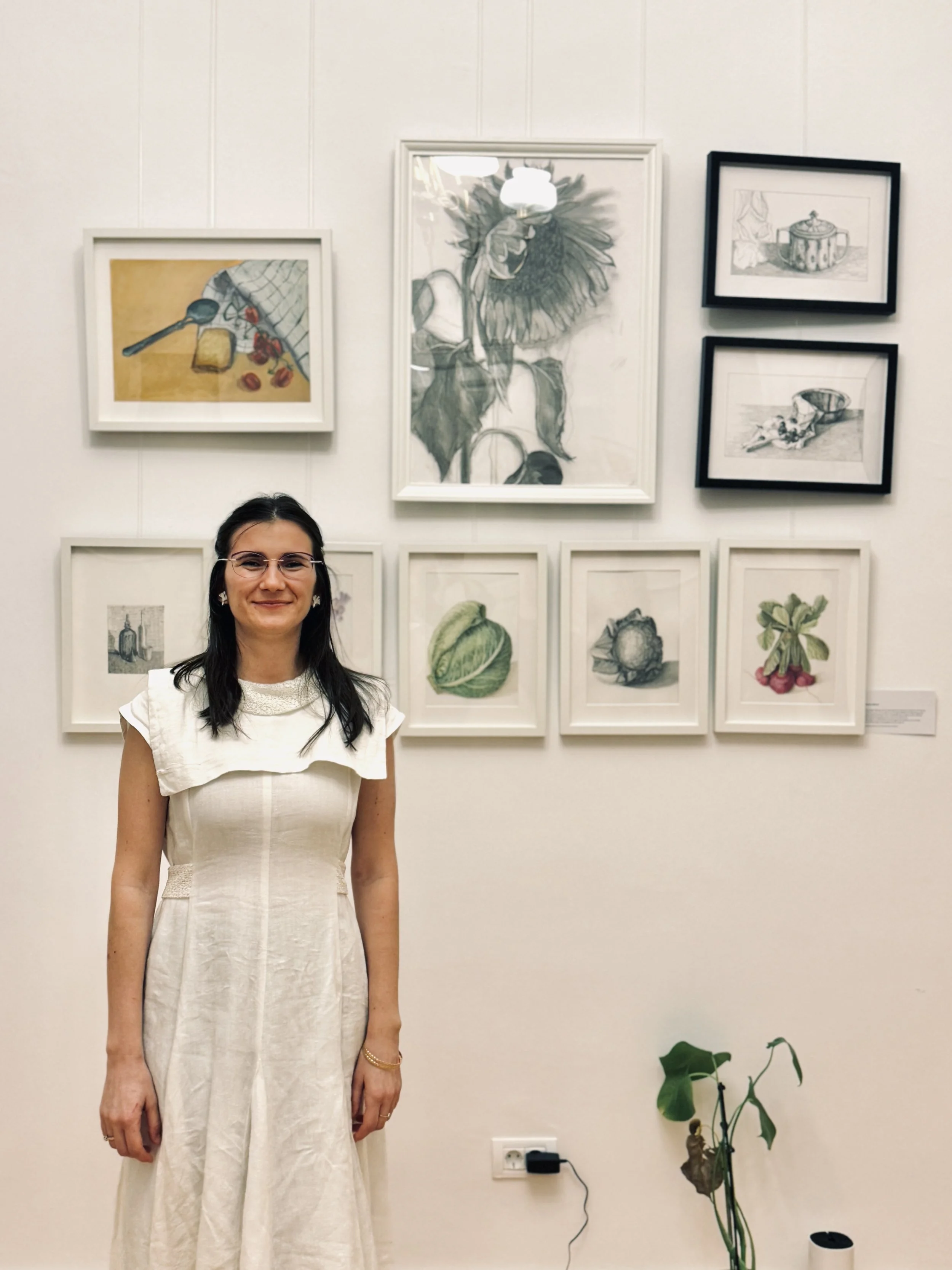 Woman in a white dress standing in front of a wall with framed art, including botanical and still life illustrations, and a potted plant on the floor.