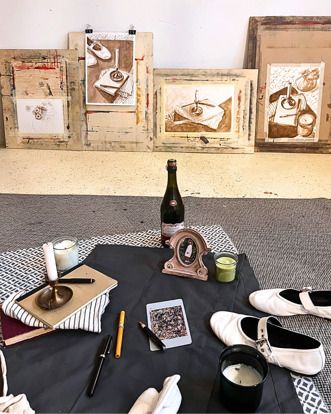 Art studio workspace with paintings in the background and a table in the foreground. The table has a black cloth, white shoes, a candle, a notebook, markers, a candle in a jar, a bottle of wine, a small framed picture, and a tablet.