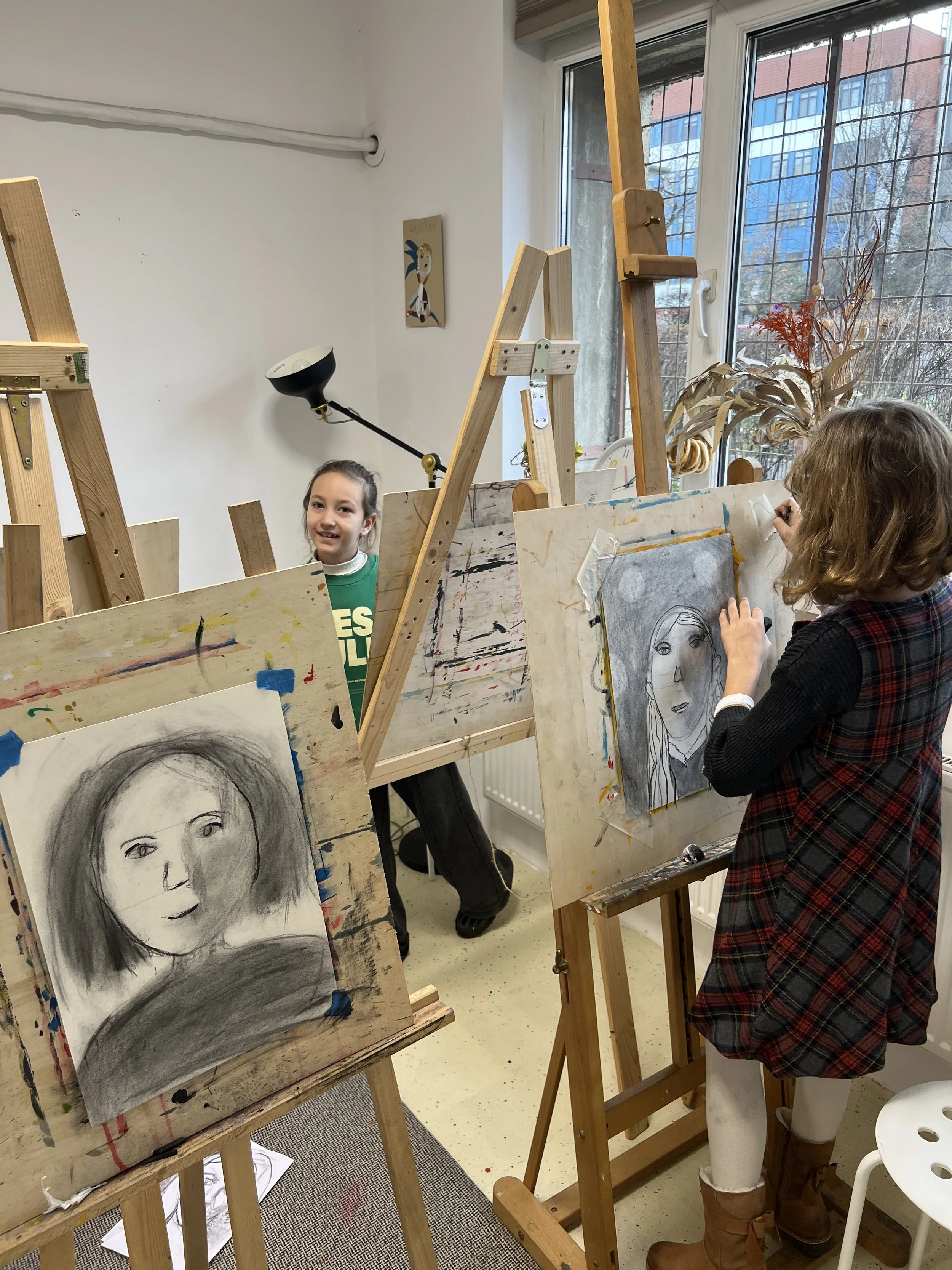 Two young girls drawing portraits on paper with charcoal in an art studio, surrounded by easels, with a large window showing trees outside.