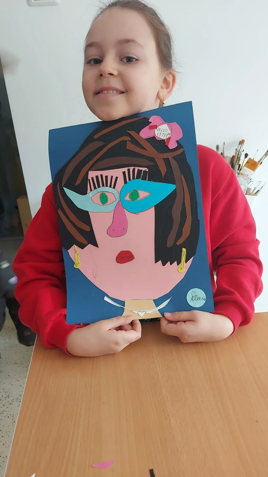 Young girl in a red sweatshirt holding a colorful abstract portrait artwork she created, with a gallery of paintbrushes in the background.
