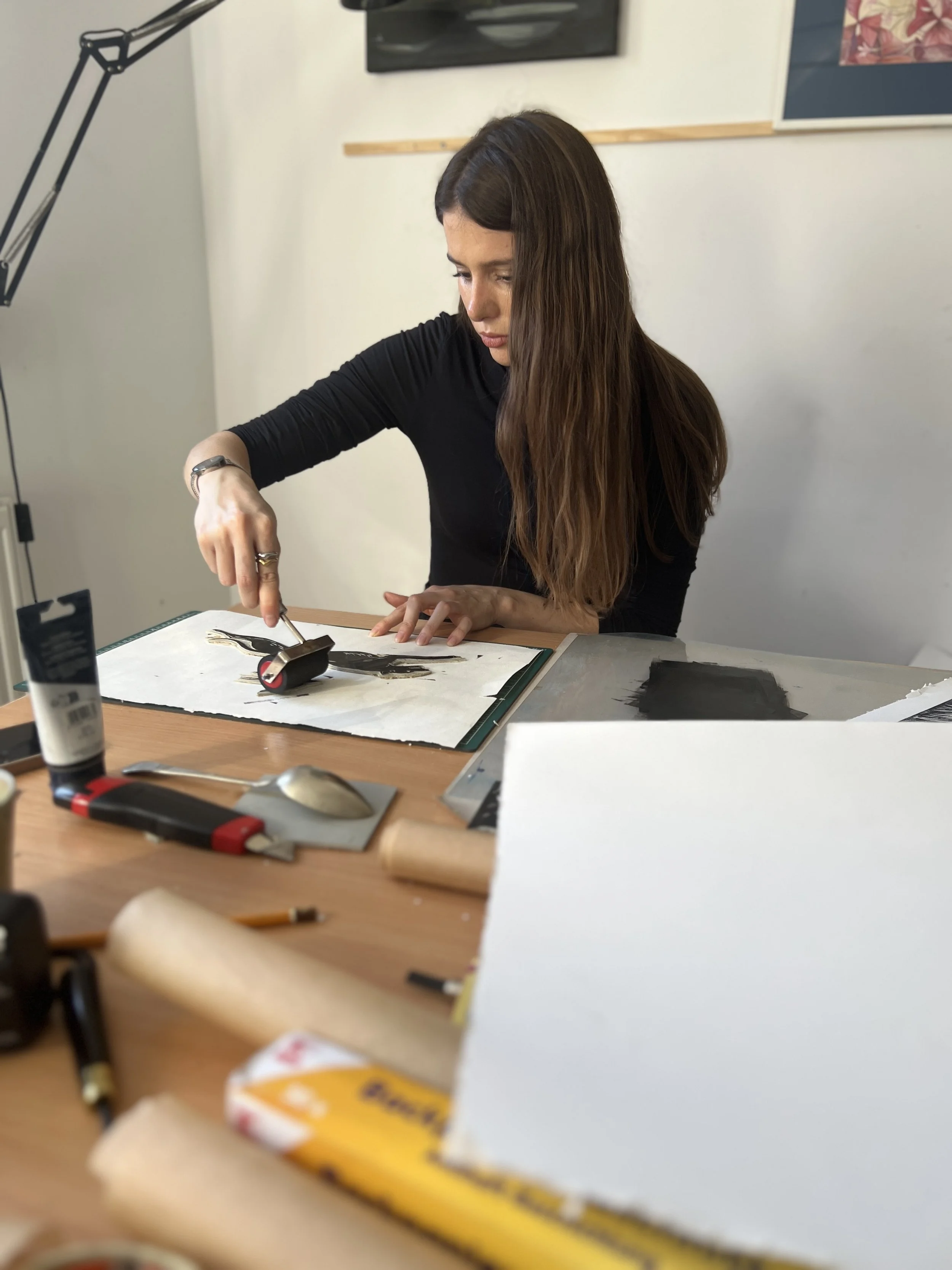 A woman with long brown hair etching an art project at a wooden table, using a tool on a piece of black and white artwork, with art supplies and paper scattered around.