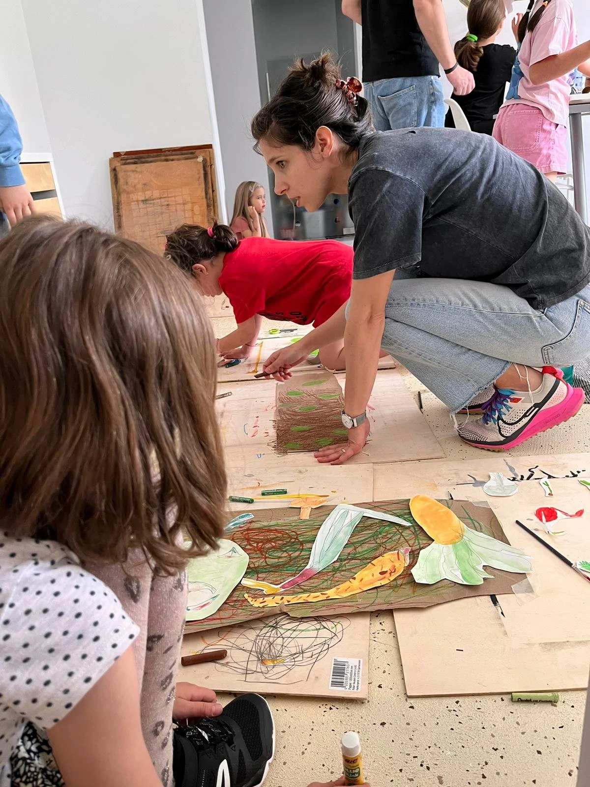 Children and an adult engaging in a creative outdoor art activity, drawing and coloring on large sheets of paper placed on the ground.
