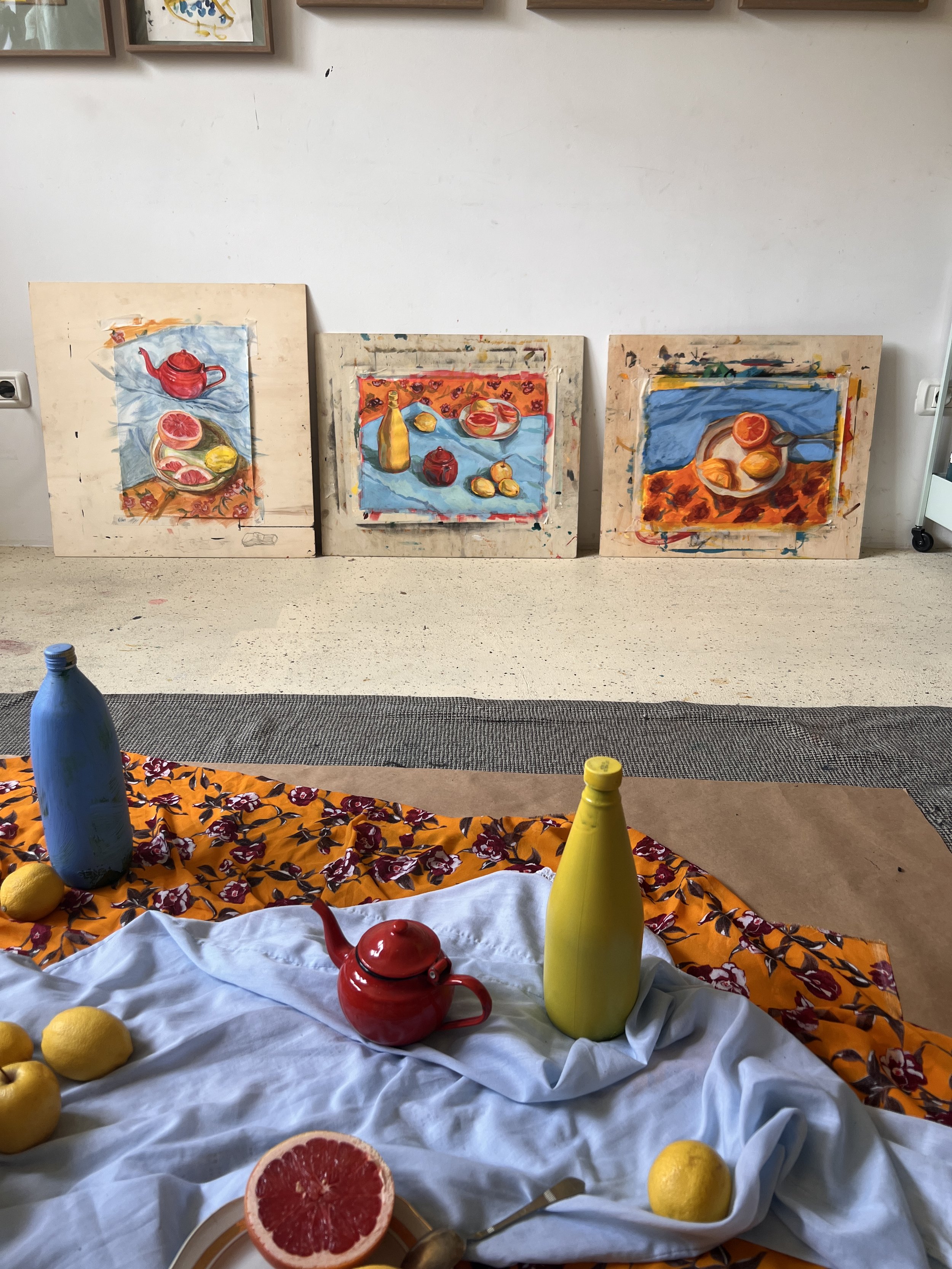 Three paintings of fruit and still life objects resting on a wall, with colored bottles, lemons, and a grapefruit on fabric in front.