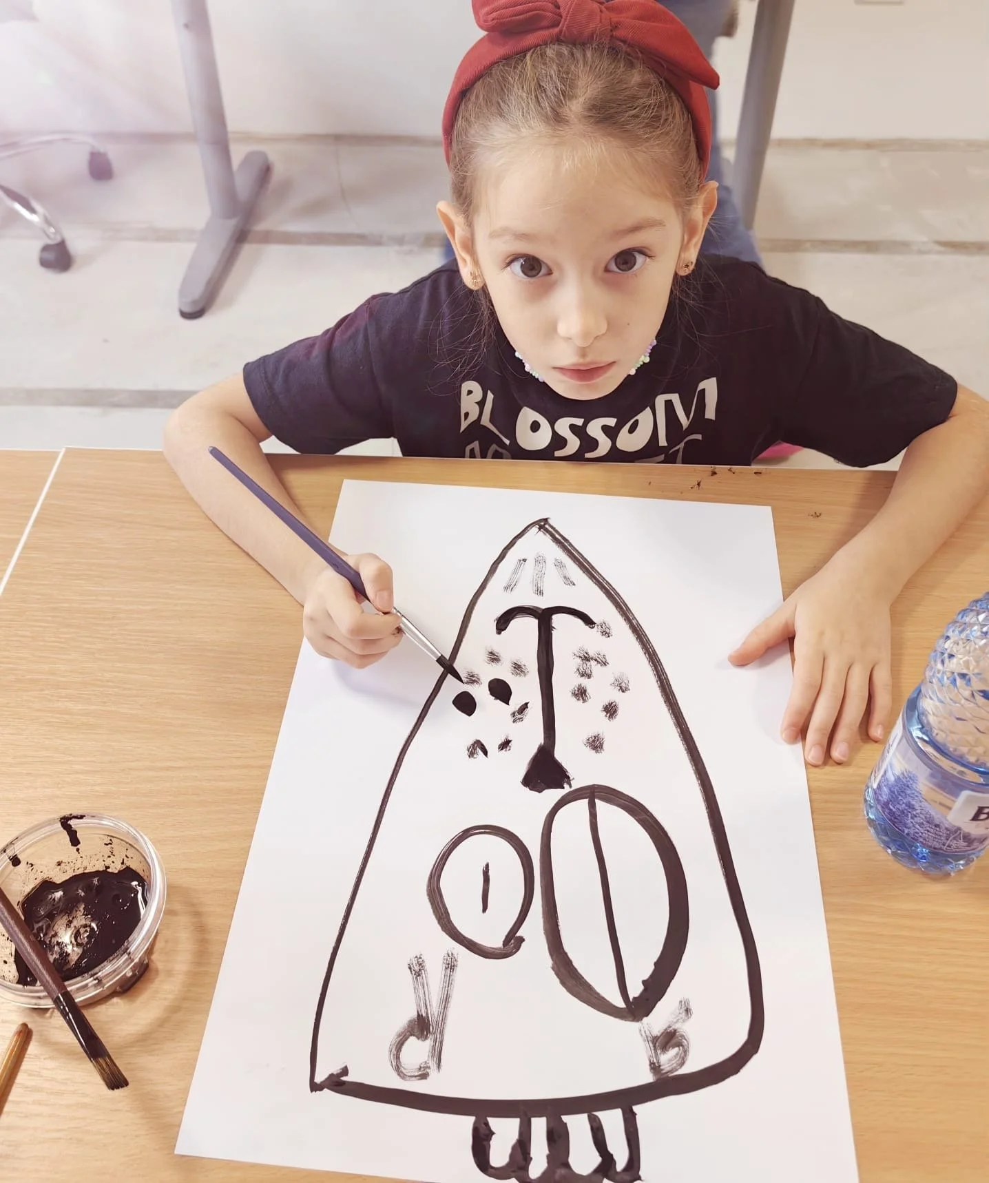 Young girl with a red headband sitting at a table, holding a paintbrush, drawing a rocket ship with black paint on a large white sheet of paper, with paint and a water bottle on the table.