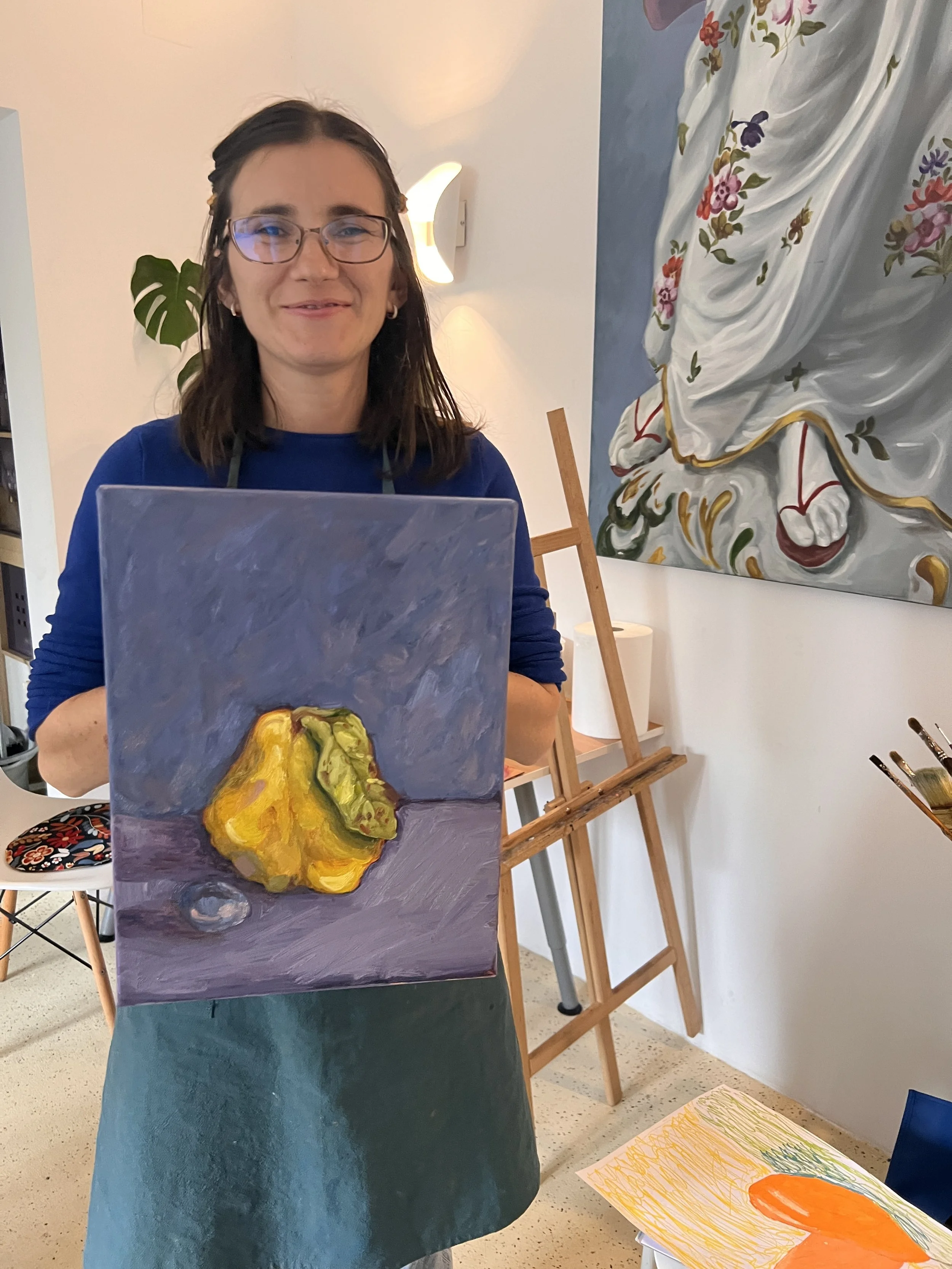 A woman with glasses and dark hair holds a painted canvas of a yellow bell pepper on a purple background in an art studio.