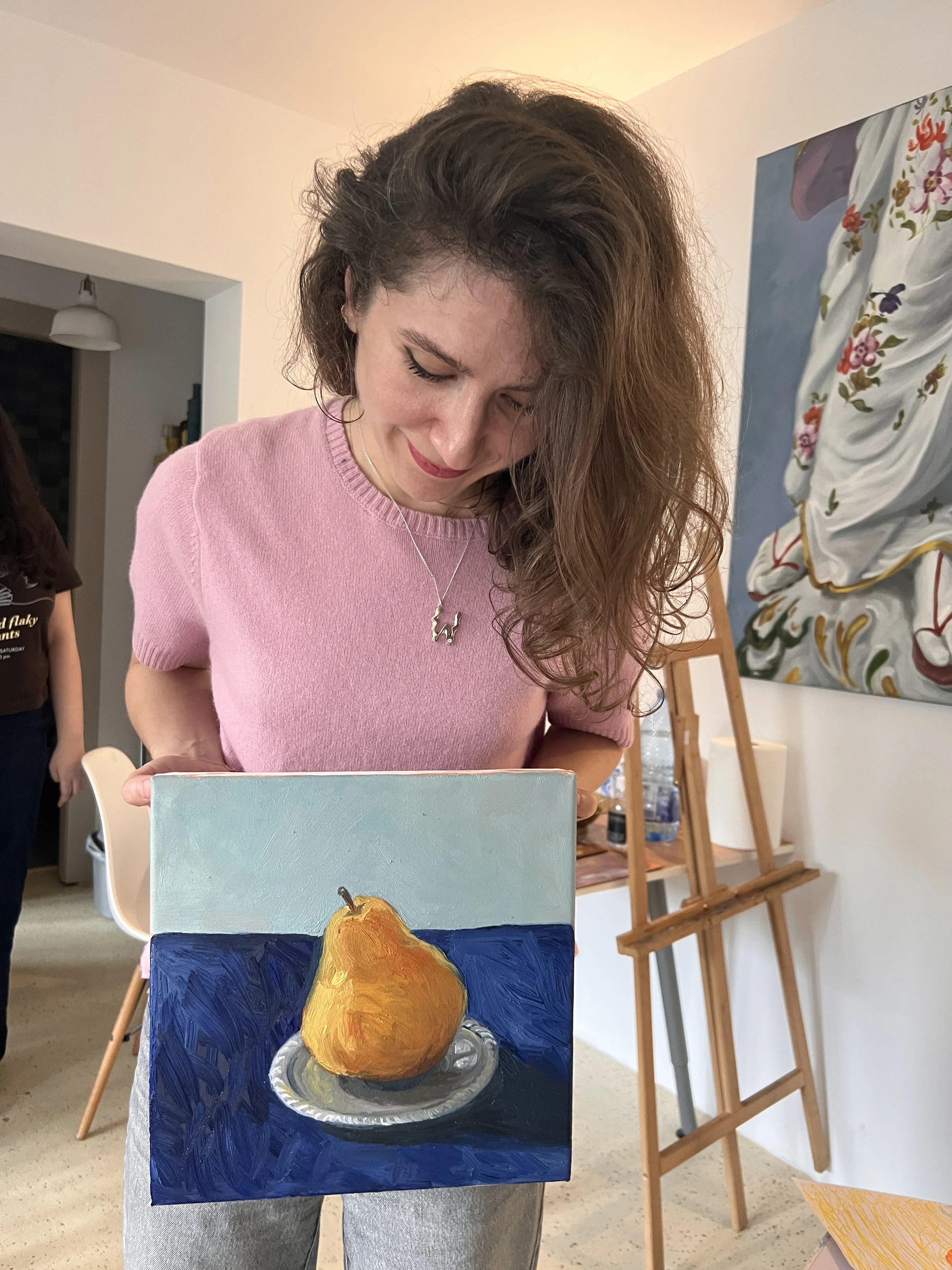 Woman in pink shirt looking down at a small painting of a pear on a plate, displayed on her stomach.