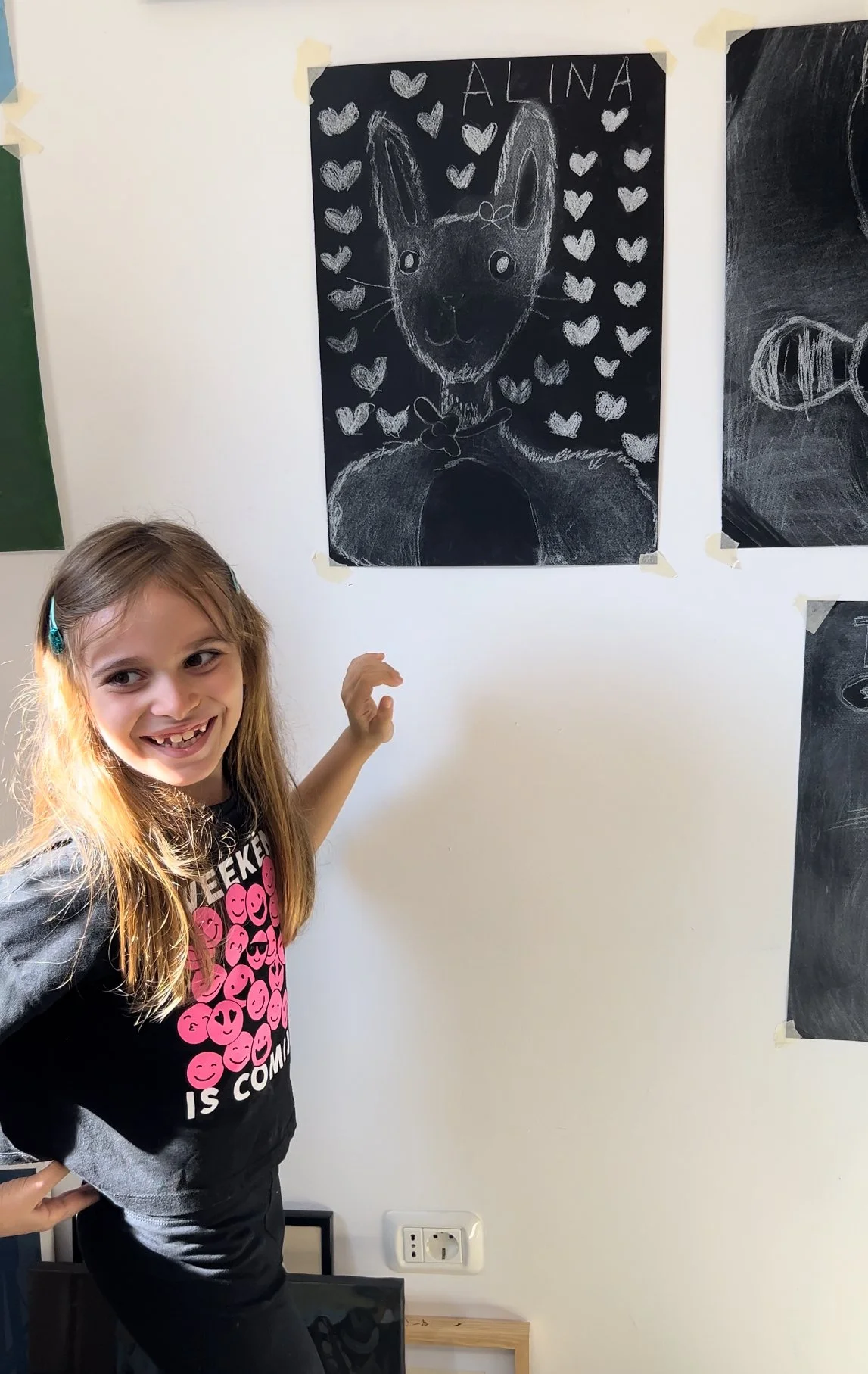 Young girl smiling and pointing at a chalkboard drawing of a cat with a background of hearts, labeled 'ALINA'.
