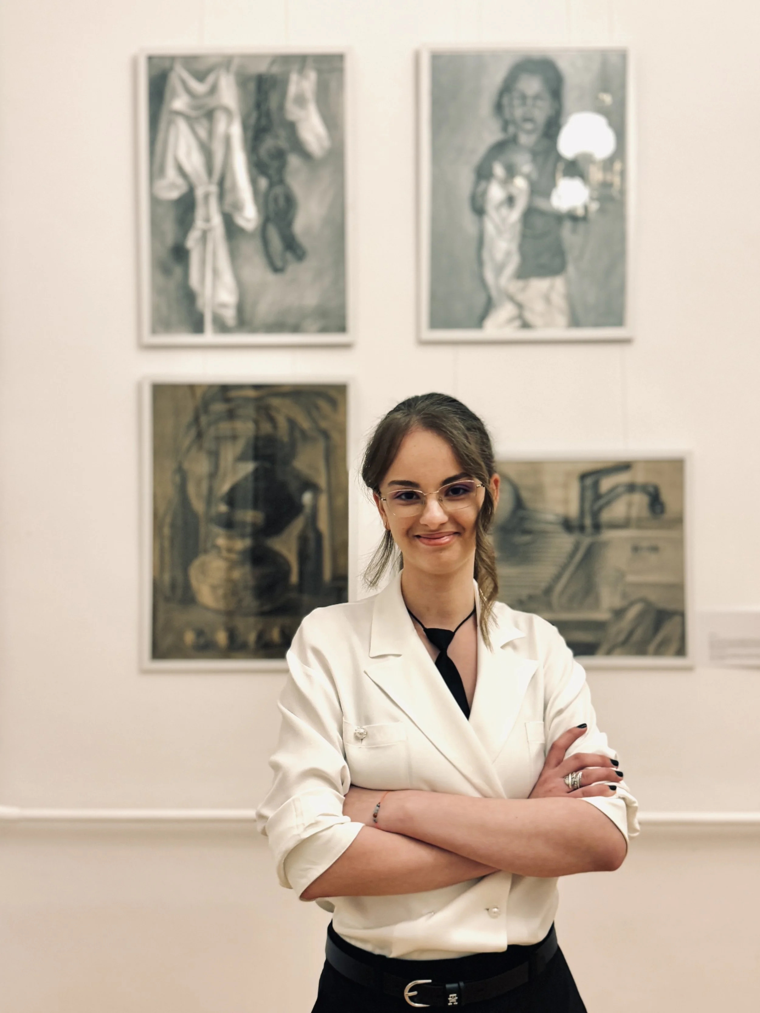 A young woman with glasses and a white shirt standing in an art gallery with four abstract paintings on the wall behind her.