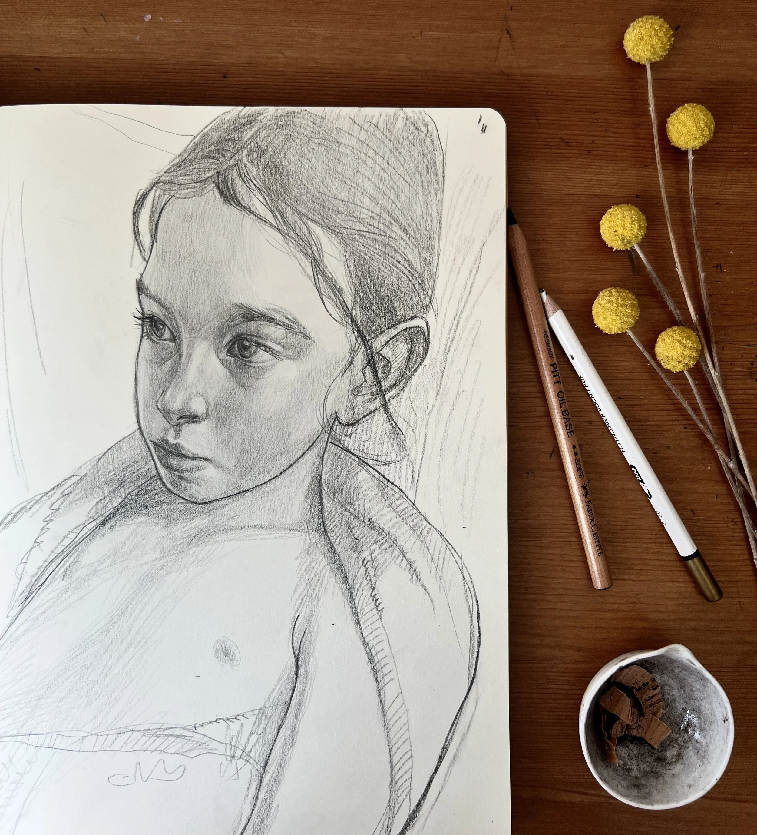 Pencil sketch of a young girl’s face and upper body on a sketchpad, placed on a wooden surface with art supplies and yellow billy ball flowers nearby.