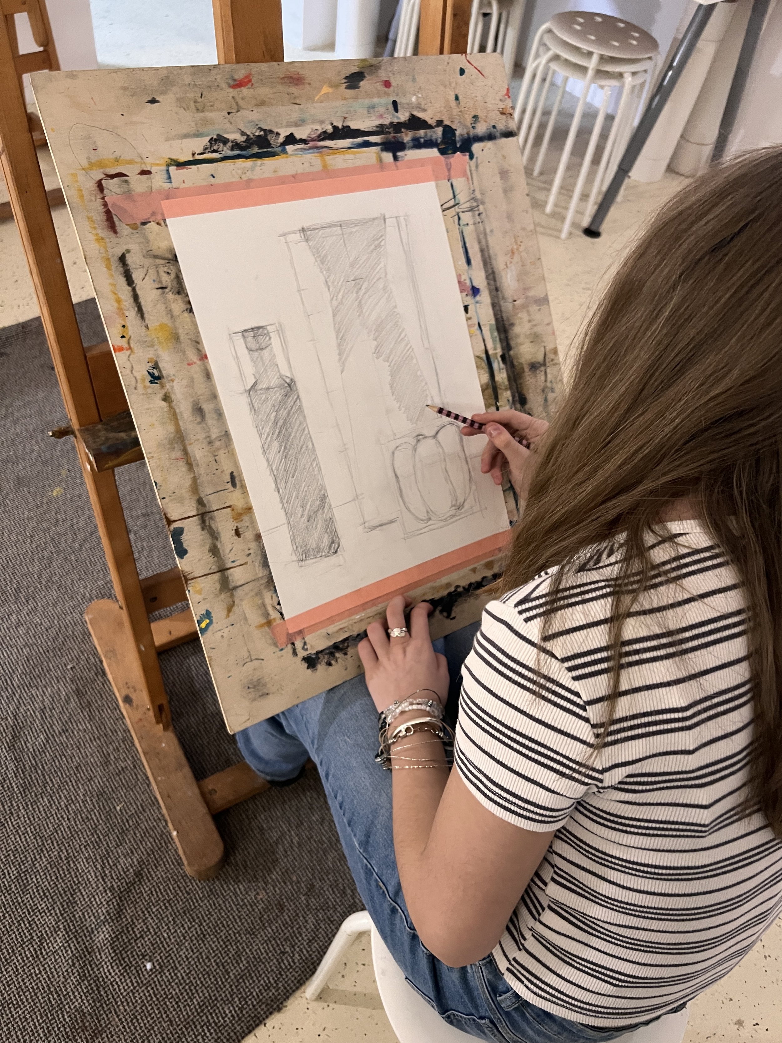 A woman drawing still life sketches of vases and bottles on paper at an art easel in an art studio.
