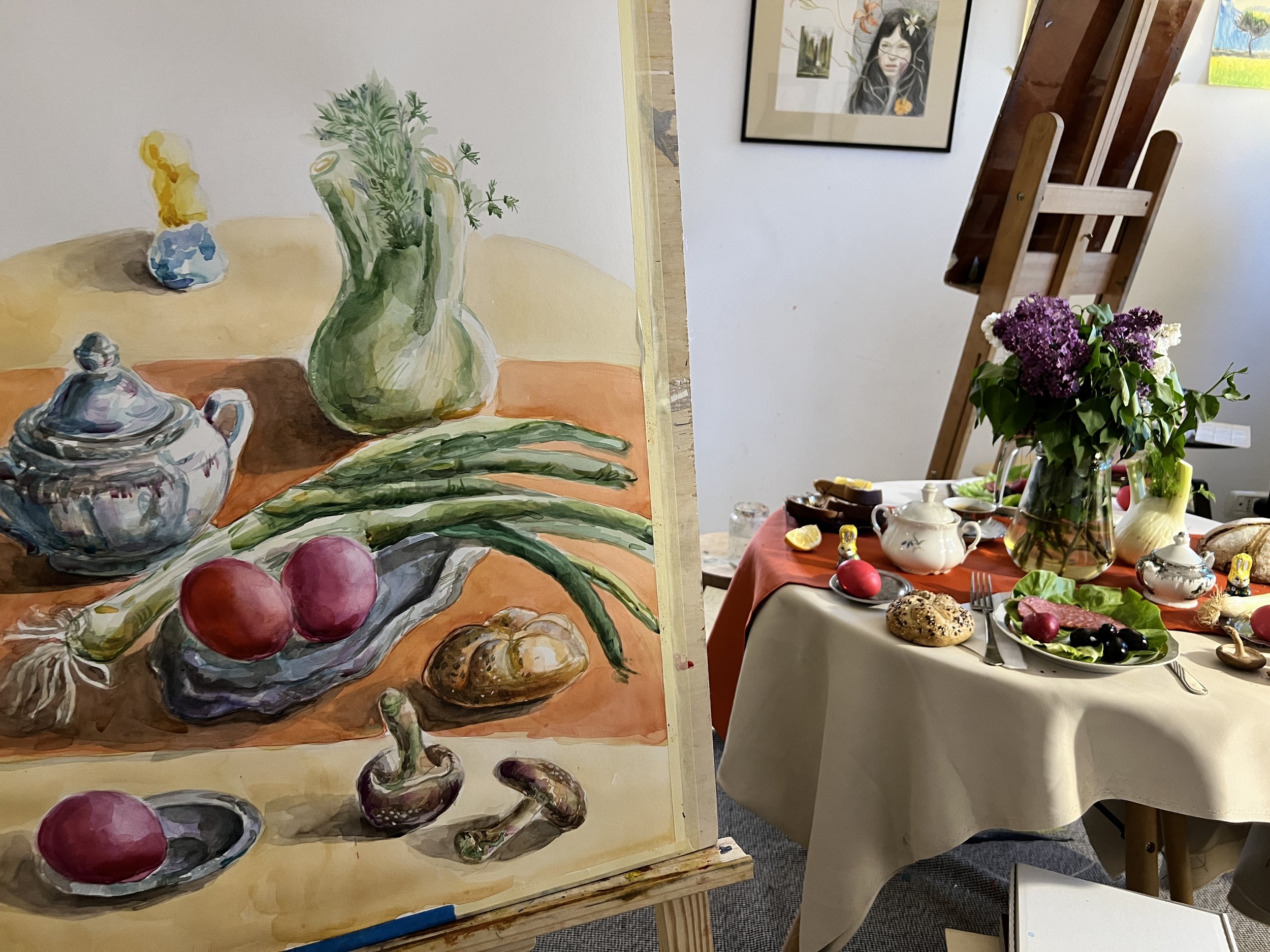 A watercolor painting of a still life with a teapot, a lid, a garlic bulb, two radishes, green beans, a leek, and a green leafy plant in a vase. The painting is placed on an easel in front of a table set with a large glass vase of purple and white fl