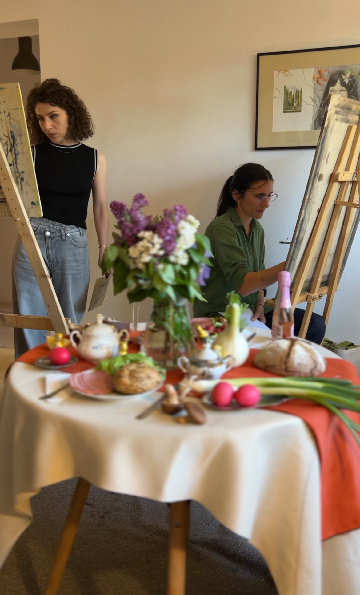 Two women painting at easels in a room with a table in the foreground decorated with flowers, bread, eggs, and a teapot.