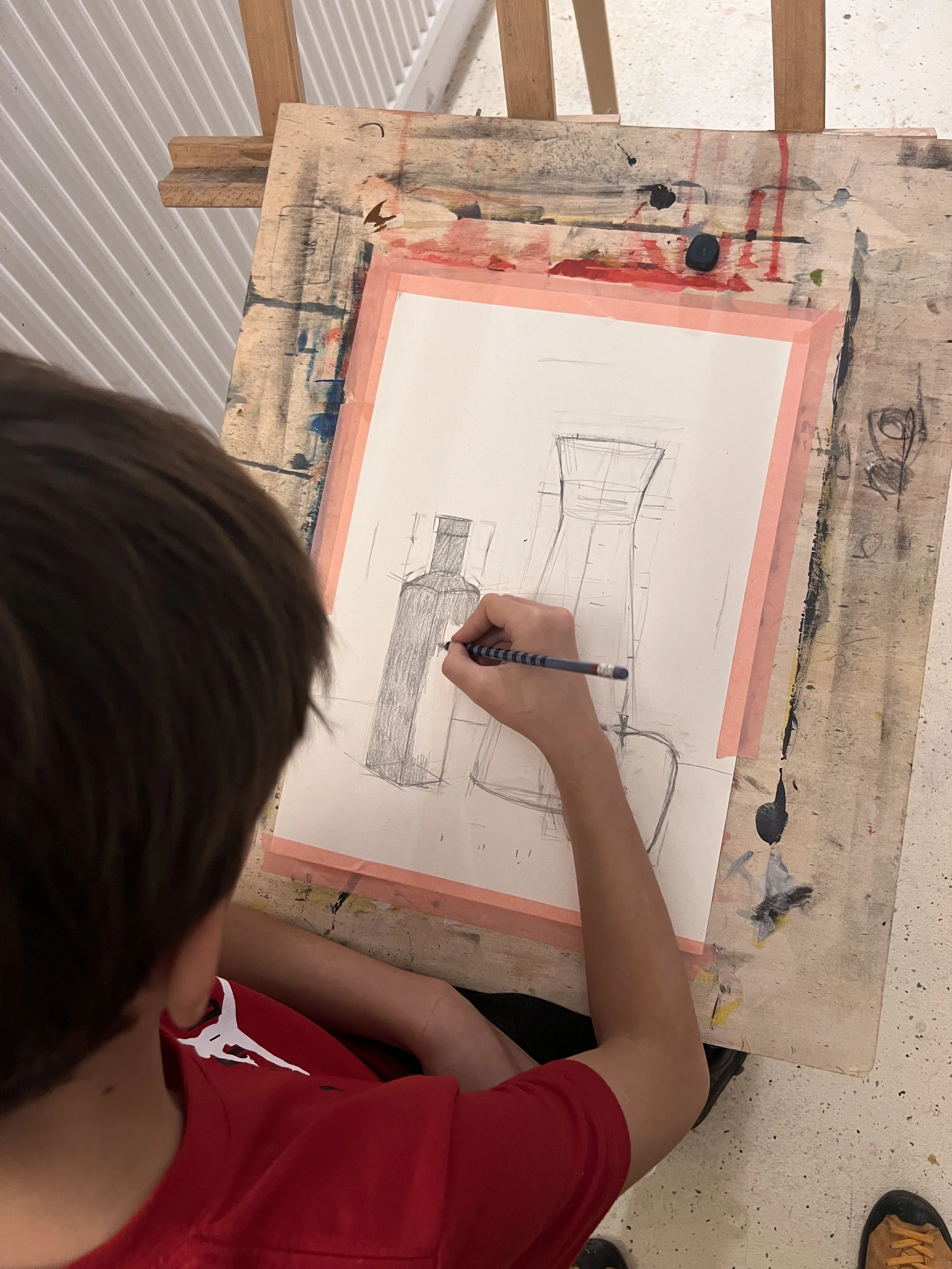 Child drawing still life sketches of bottles on a sheet of paper, at a cluttered art workspace.