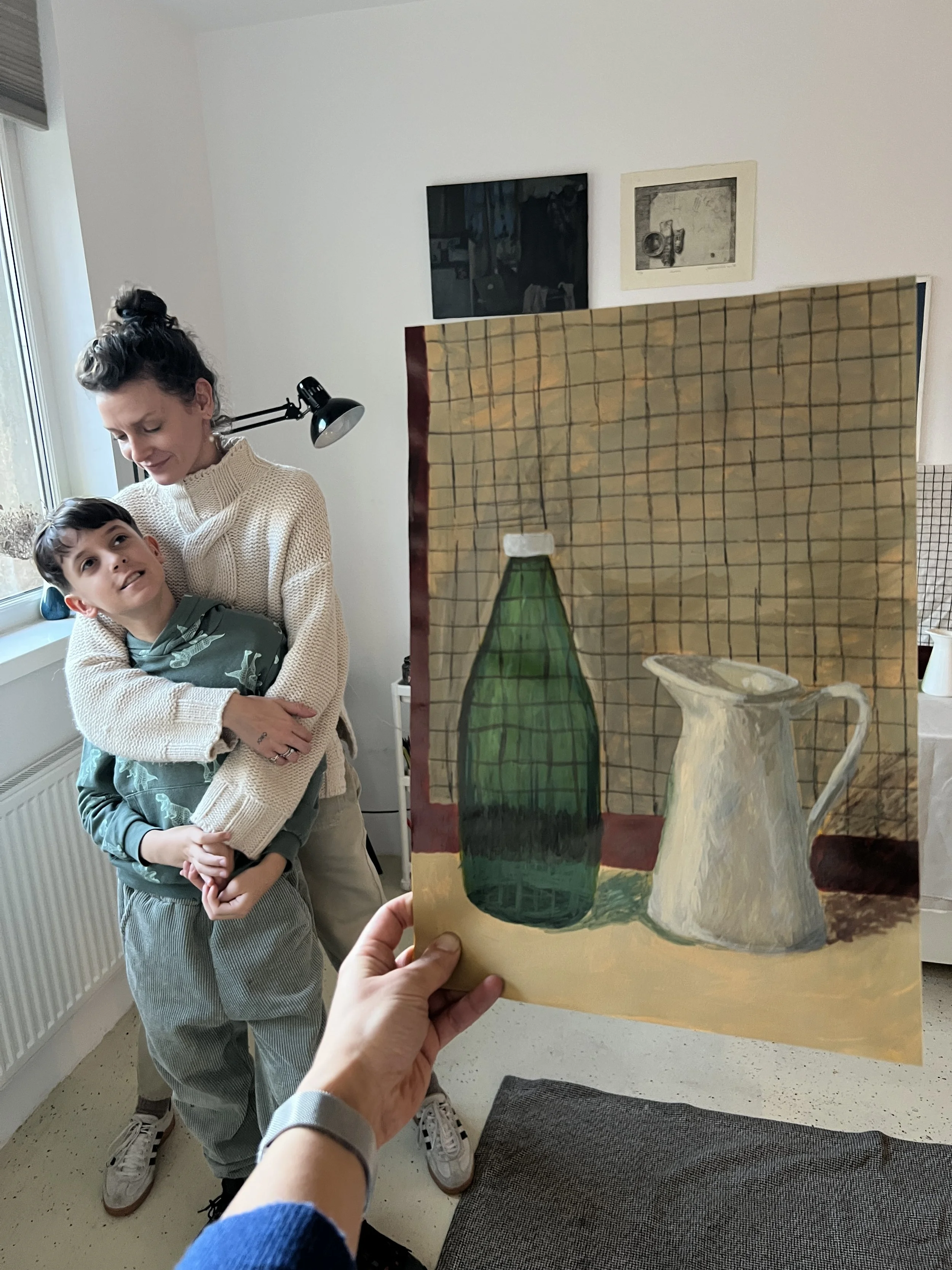 A person holding a painting of a green bottle and a cream pitcher in front of a woman and a young boy in a room with white walls, artwork, and a window.