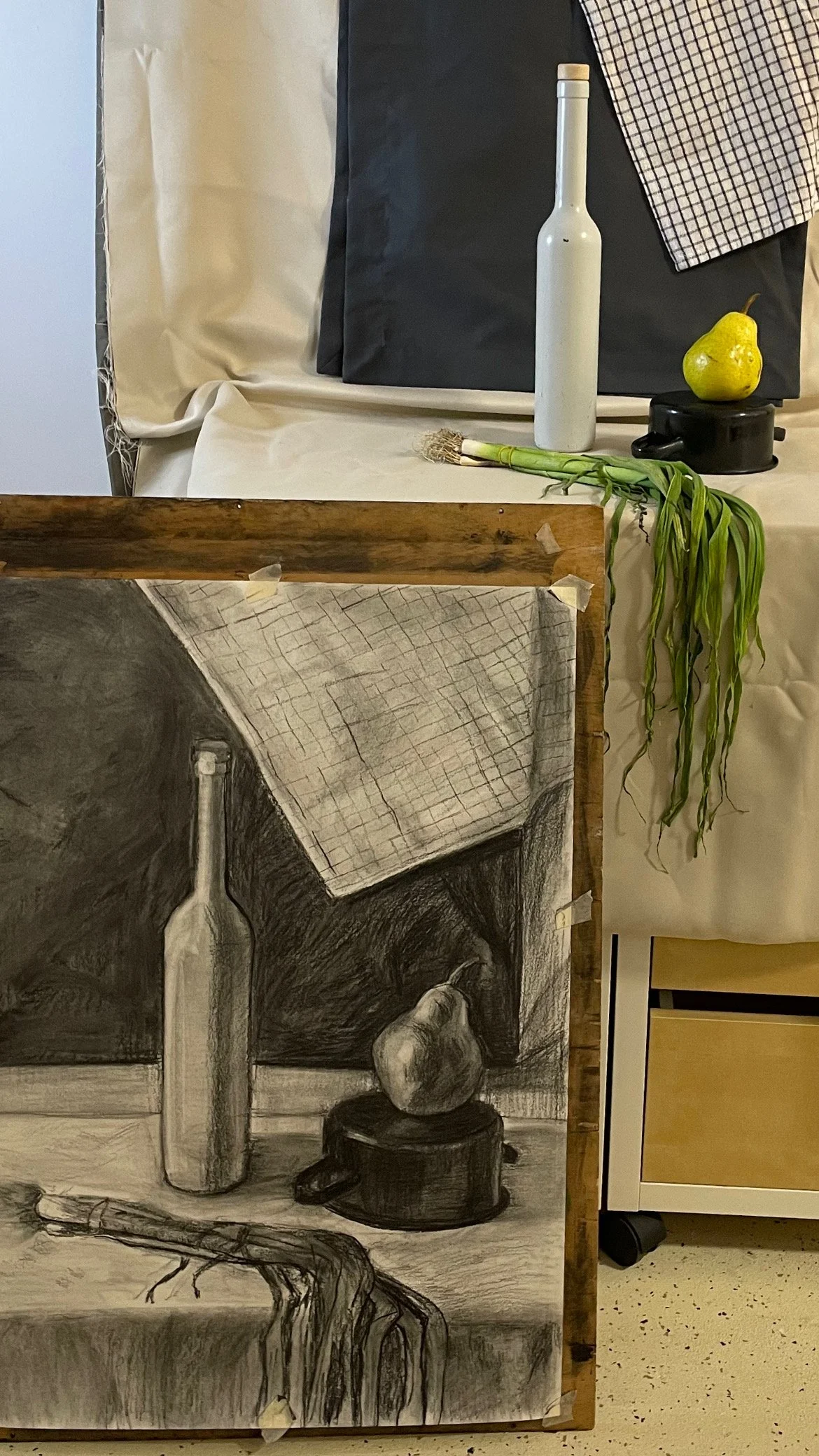 A still life arrangement featuring a painted black and white still life on a wooden board, a real white bottle, a yellow pear, a bunch of green onion, and a black object on a table with fabric and paper background.