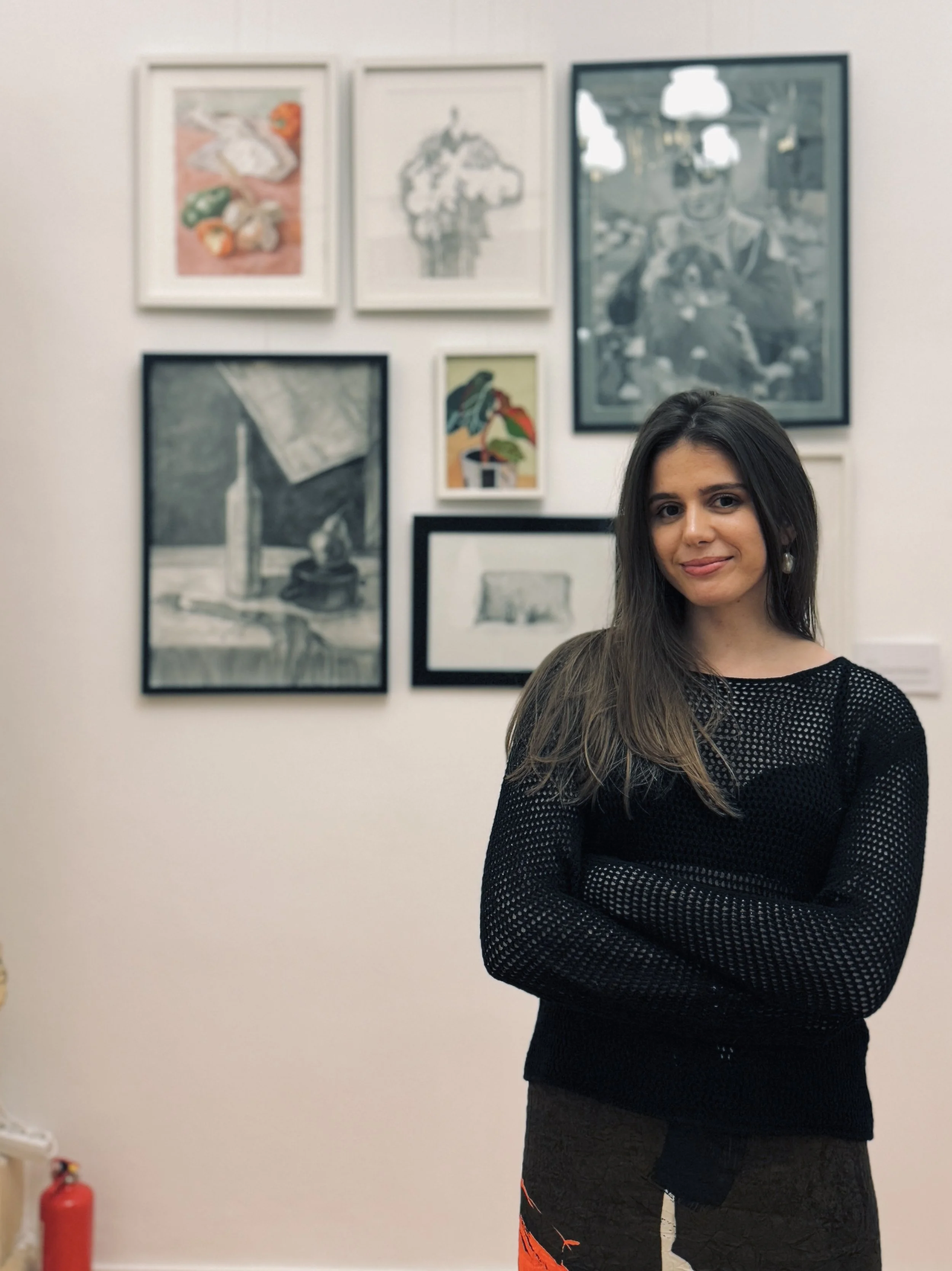 A young woman with long dark hair wearing a black perforated sweater standing with arms crossed in front of an art gallery wall with various framed artworks.