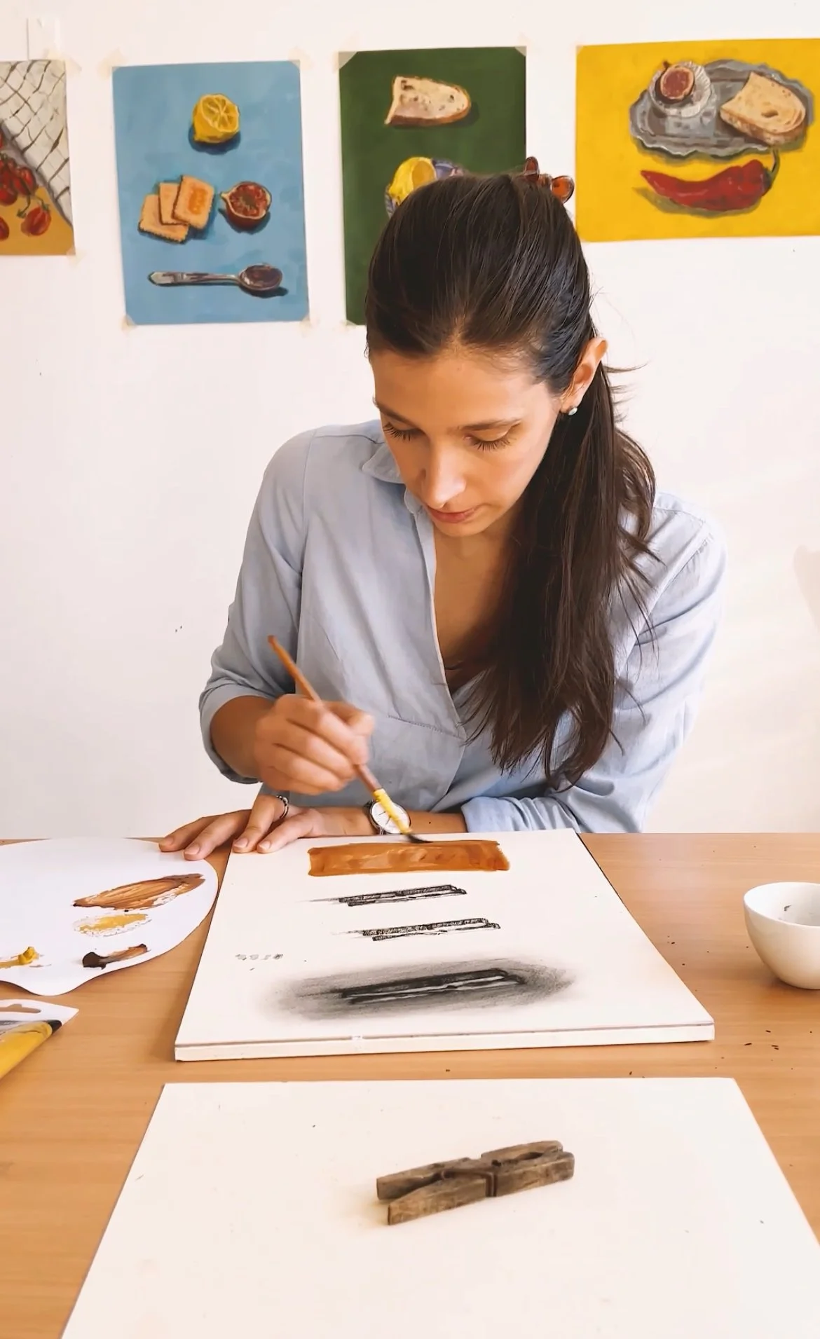 A woman painting abstract art on a white canvas, sitting at a wooden table, with several paintings of food images on the wall behind her.