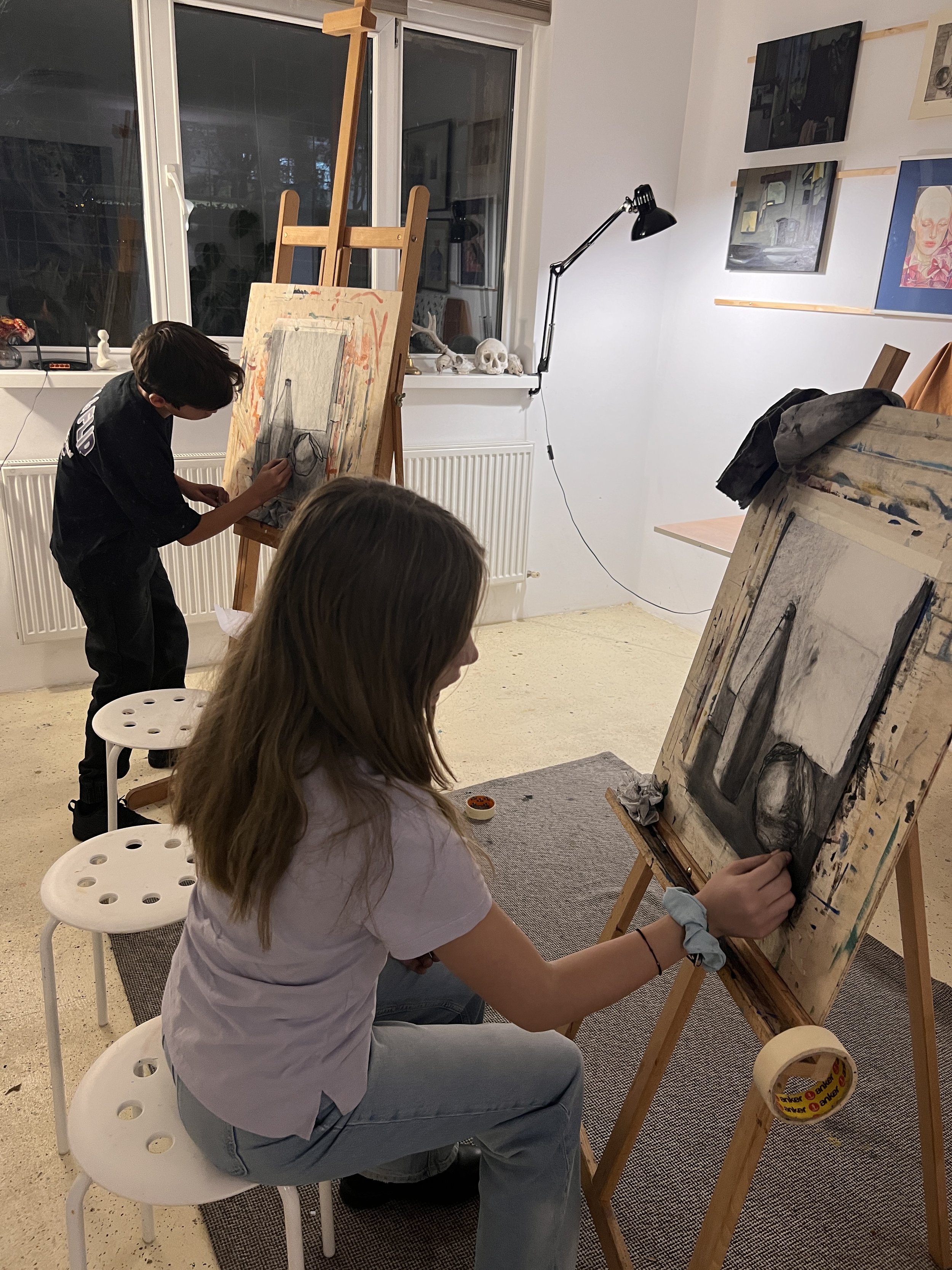 Two children, a boy and a girl, are painting on canvases set up on easels in an art room. The boy, standing, is working on a still life painting, while the girl, sitting on a white stool, is focused on her artwork. The room has artwork on the walls, 