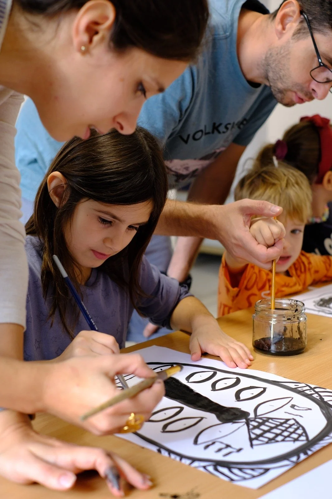 Children and adults doing a painting activity together at a table, with a paper featuring a large leaf and fruit design, some of the children are using string and paint.