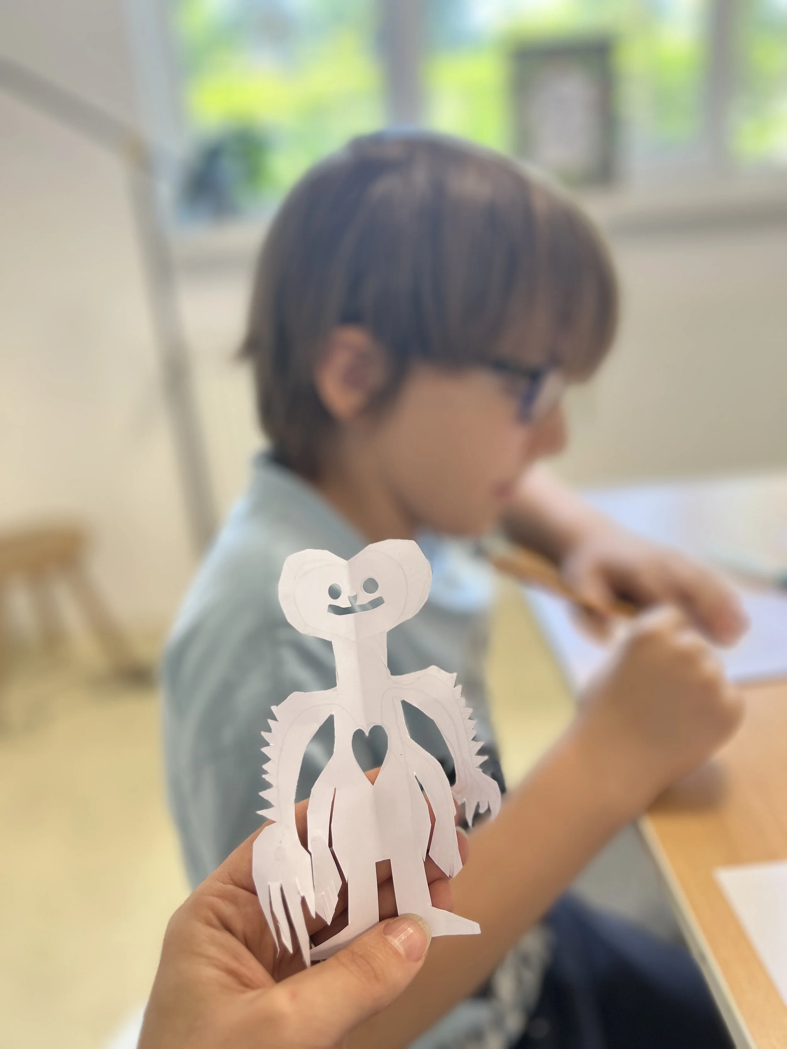 A paper craft of a smiling creature with a heart-shaped cutout on its chest, held in front of a boy drawing at a desk in a classroom.