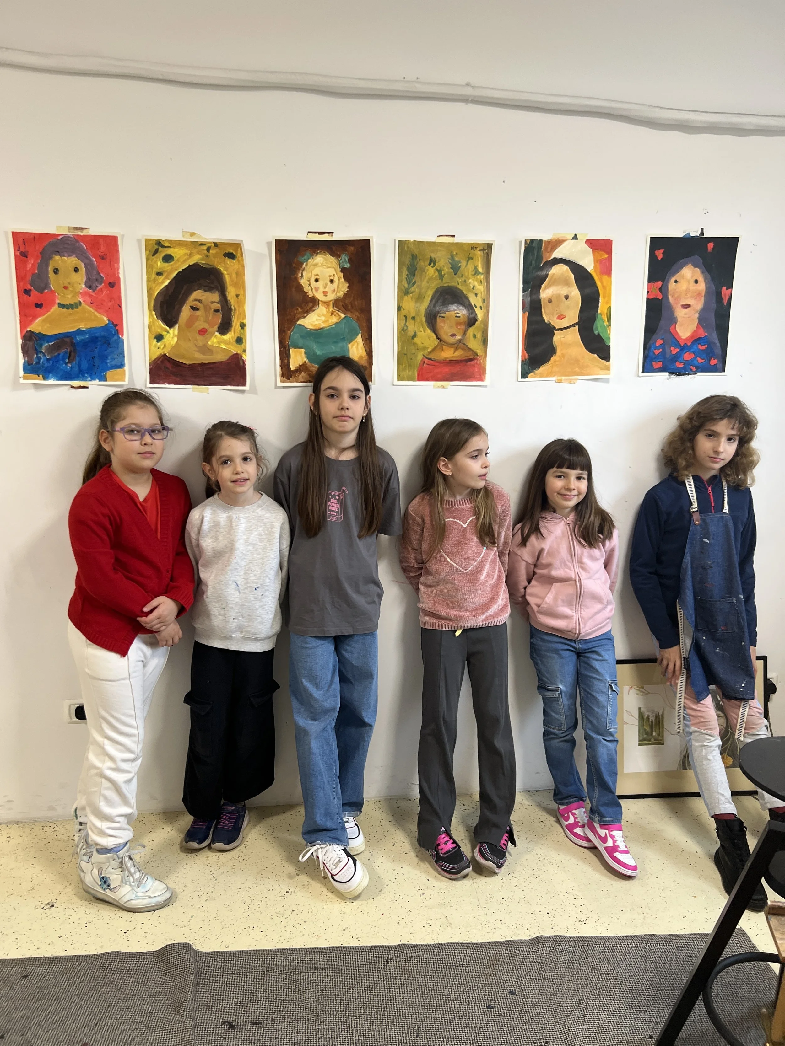 Six young girls stand in front of a wall displaying six colorful painted portraits of women, possibly self-portraits or inspired by famous figures, in an art gallery or classroom.