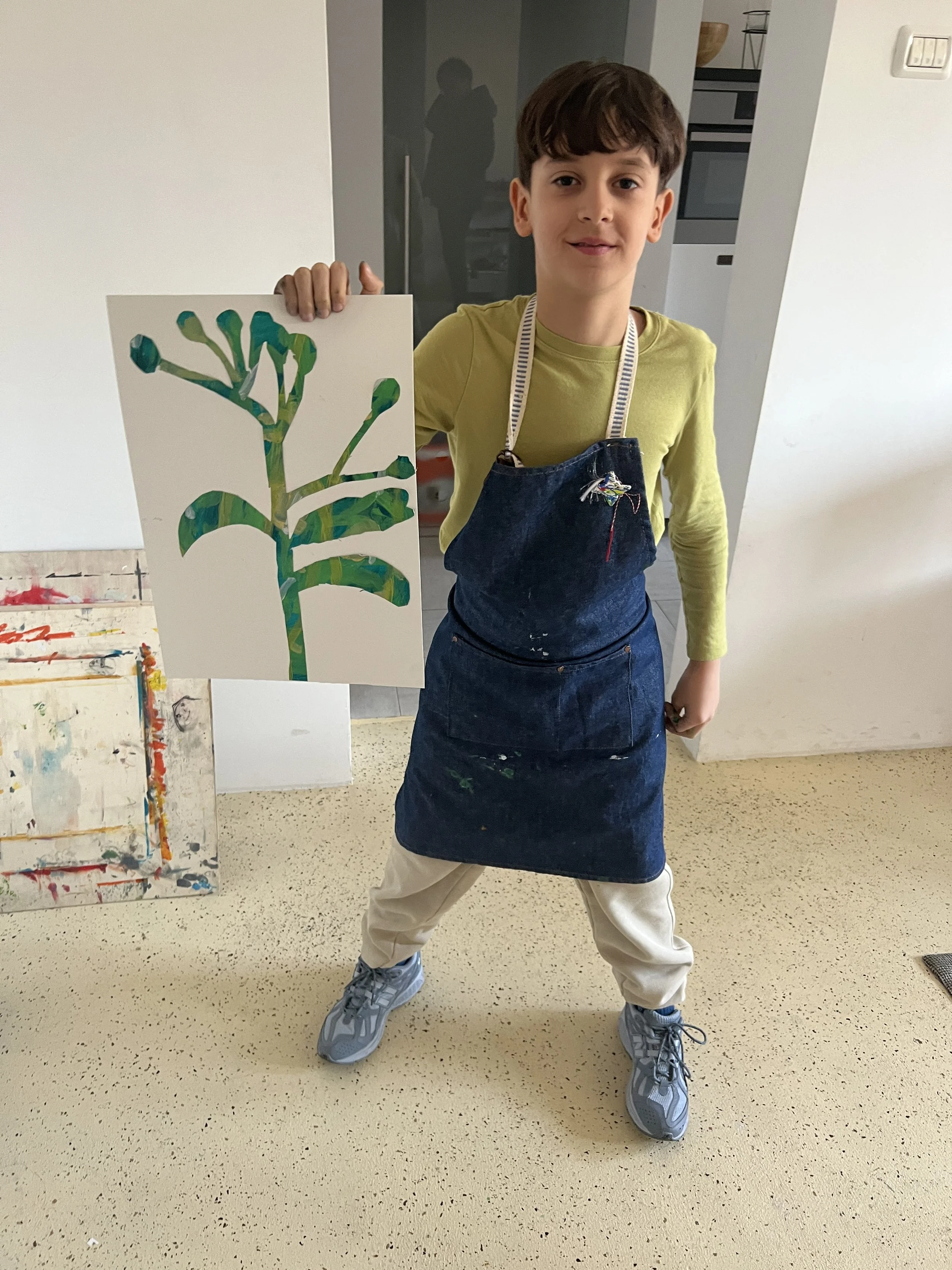 A boy in a yellow shirt and blue apron is standing on a beige floor, holding a colorful painting of a plant or tree.