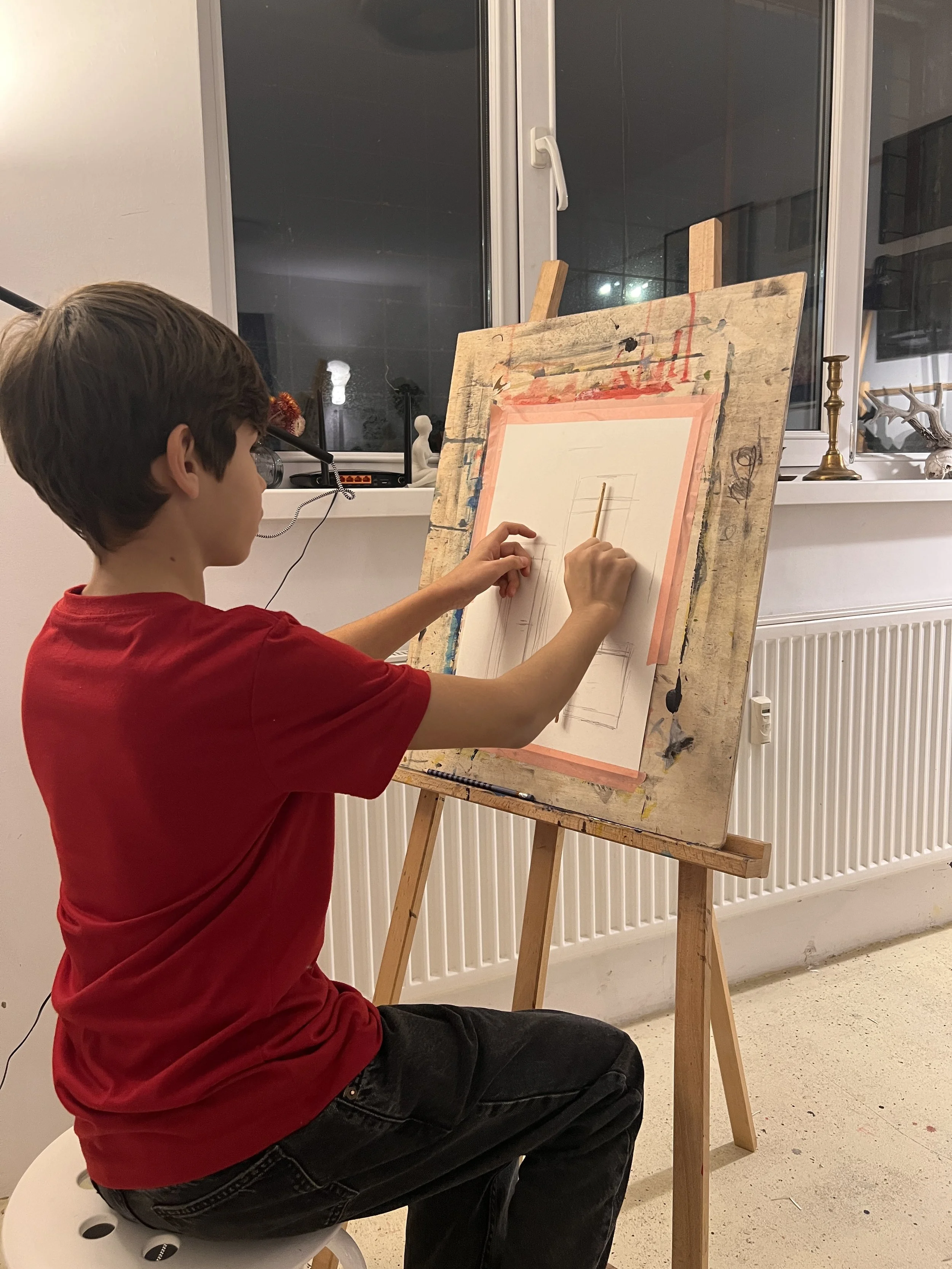 A young boy in a red shirt sitting on a white stool, drawing with a pencil on a large sheet of paper mounted on an easel in a room with a window and some decorative objects on the windowsill.