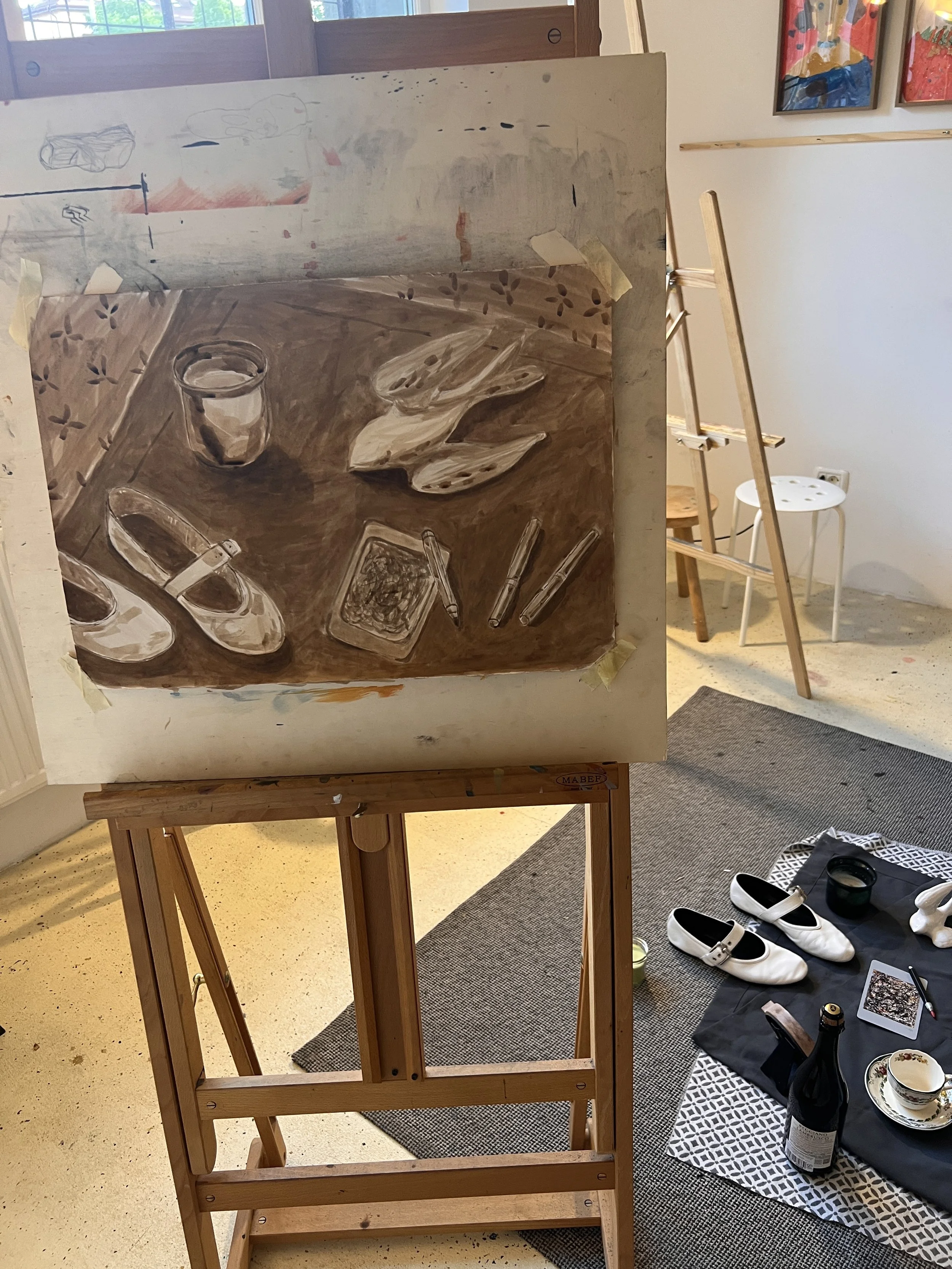 A still life painting on an easel shows shoes, a jar, bread, and various objects on a wooden table.