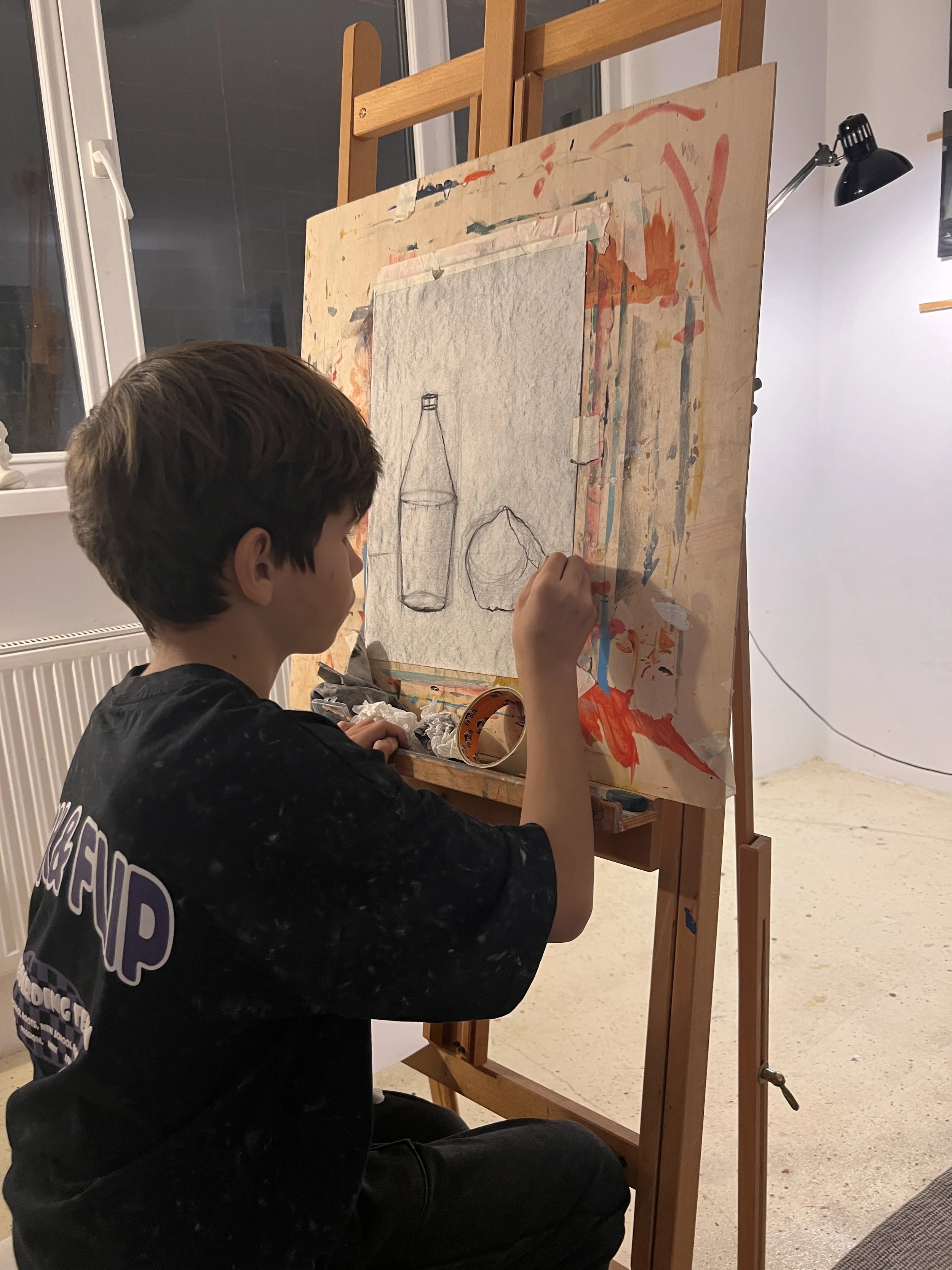 A boy sitting on the floor working on a drawing of a bottle and a sphere on an easel in a well-lit room.