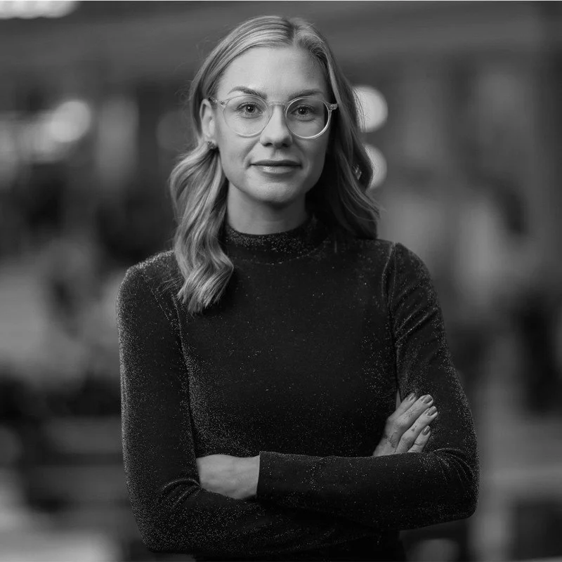 A woman with shoulder-length wavy hair and glasses standing with crossed arms, looking at the camera, in a blurred indoor setting.