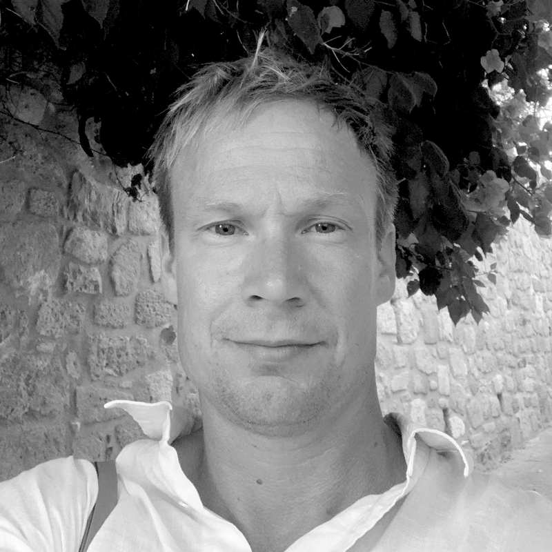 A man outdoors taking a selfie with a brick wall and leafy tree in the background.