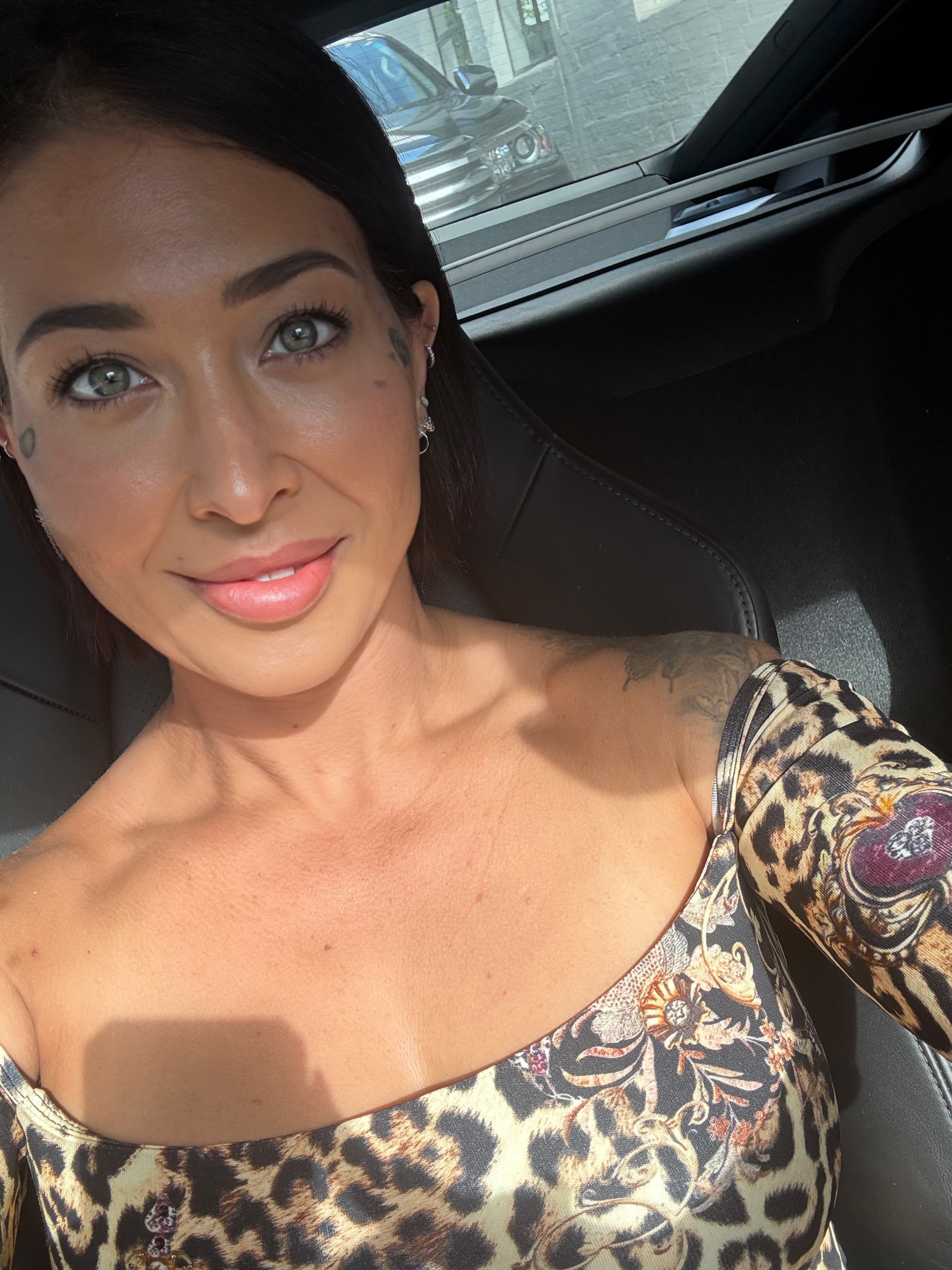 A woman with dark hair and fair skin taking a selfie inside a car, wearing a leopard print off-shoulder top, with a slight smile and blue eyes.