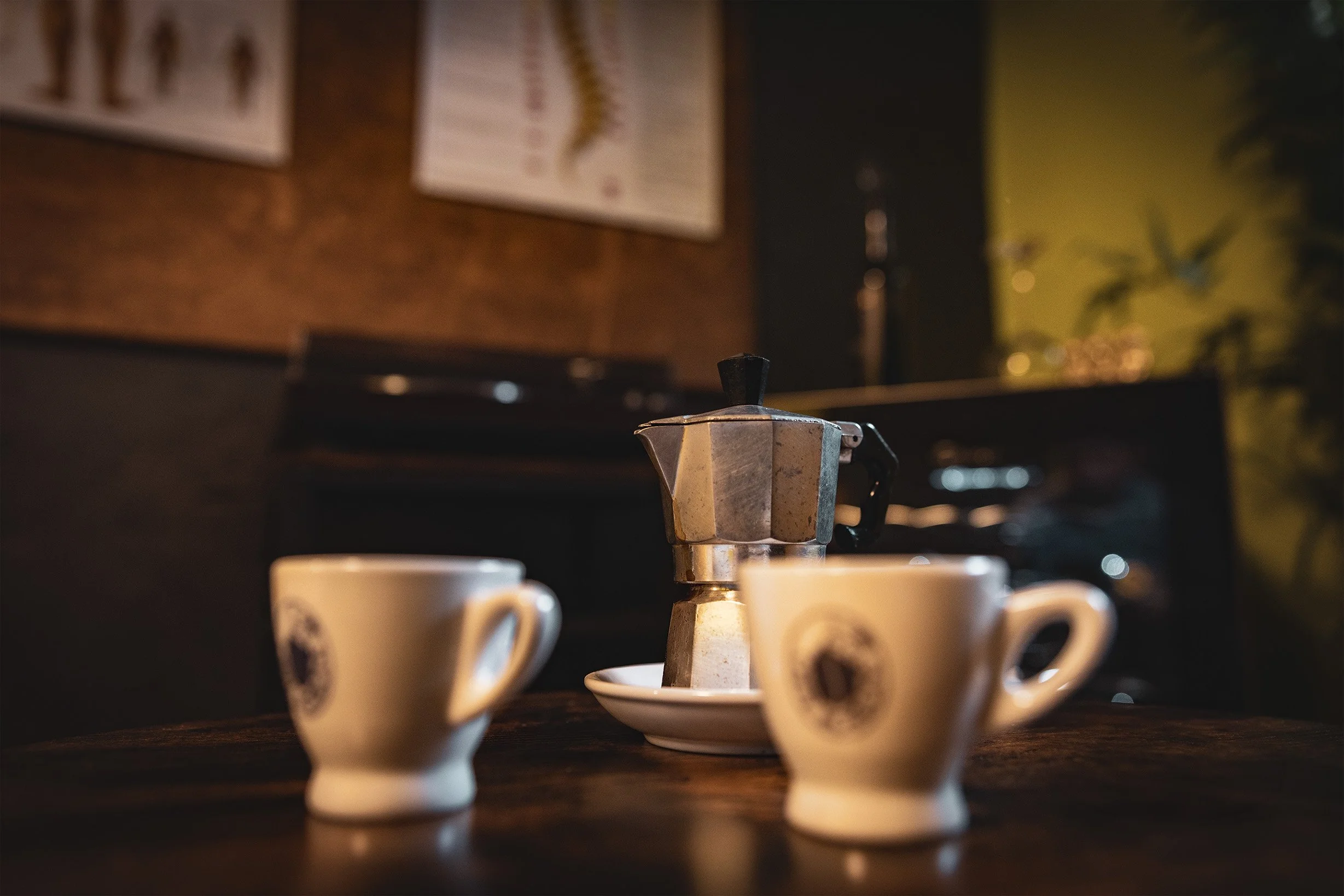 Zwei Espressotassen vor einer Moka-Kaffeemaschine auf einem Holz-Tisch in einer gemütlichen, warm beleuchteten Umgebung.