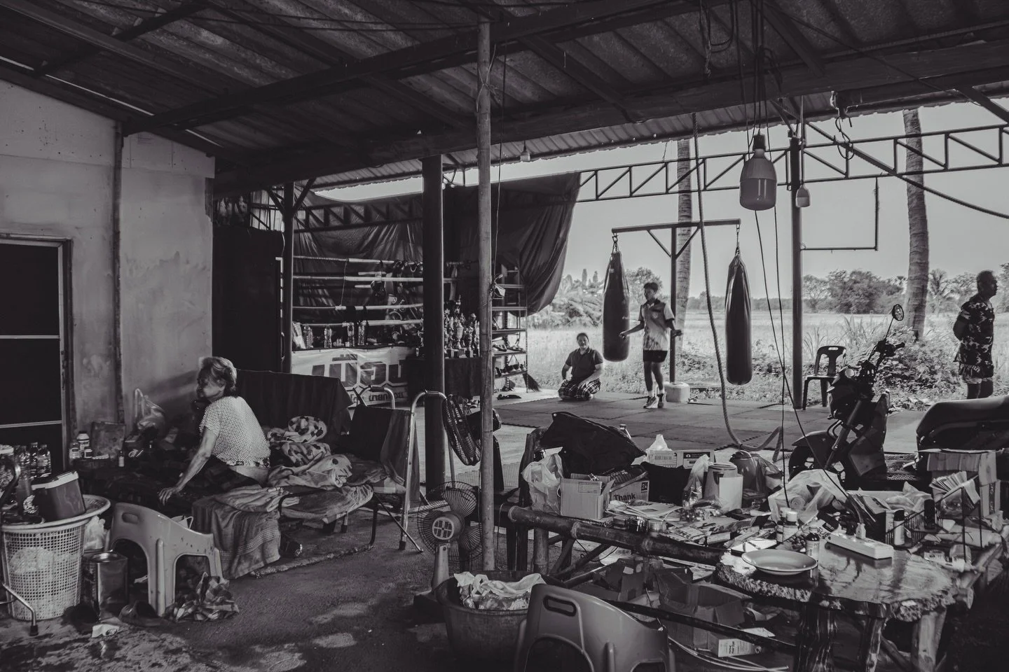 A wide view of PorMuangPhet Gym. The single ring dominates the space, flanked by 2 punching bags, 2 ropes, and a pair of tires for training. 

Just a few feet away, Lek MuangPhet&rsquo;s mother rests in a bench that serves as her bed at night. Nearby