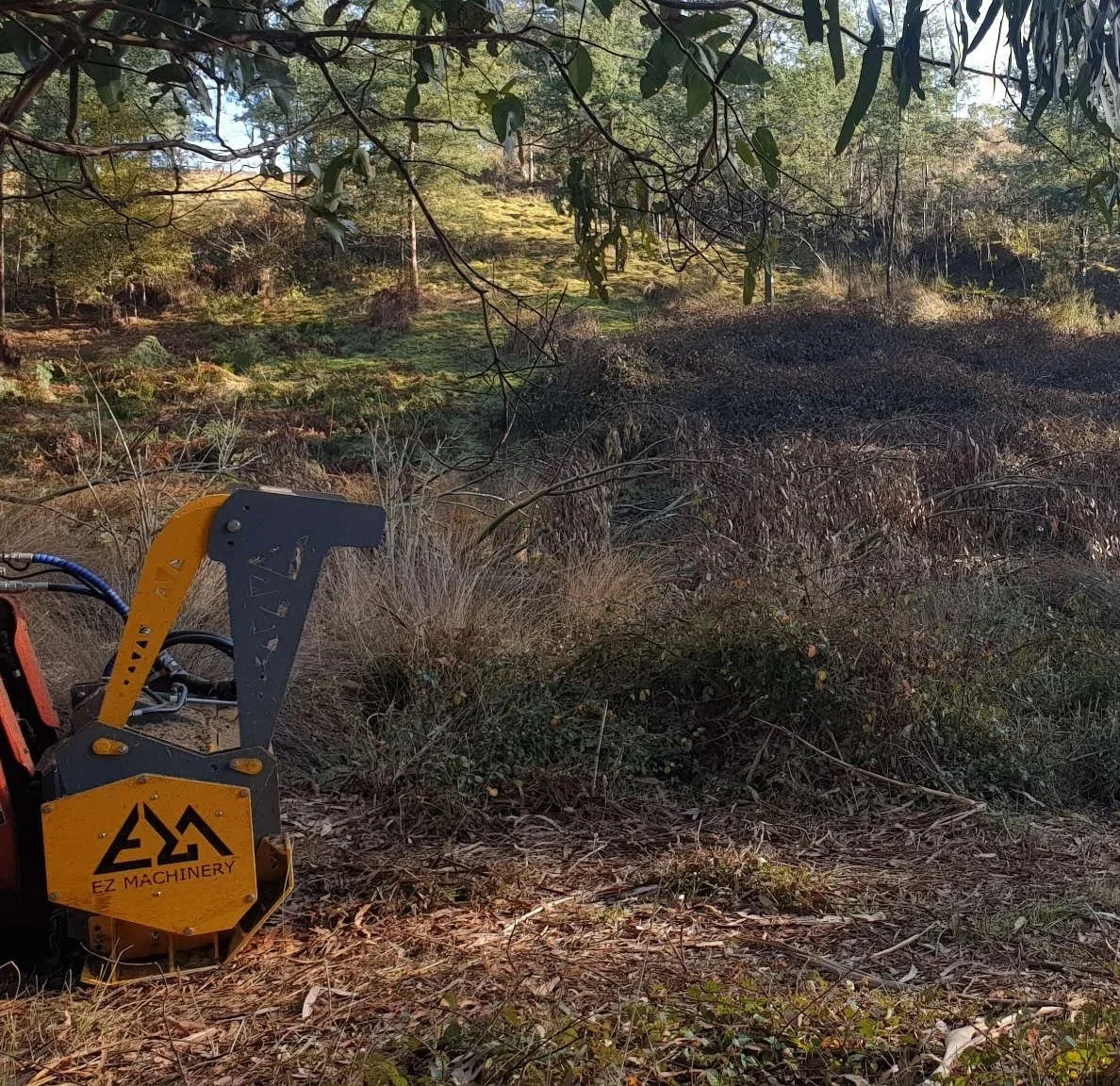 A Bush Hog rotary cutter with yellow and gray parts in a wooded area, with trees and bushes in the background.