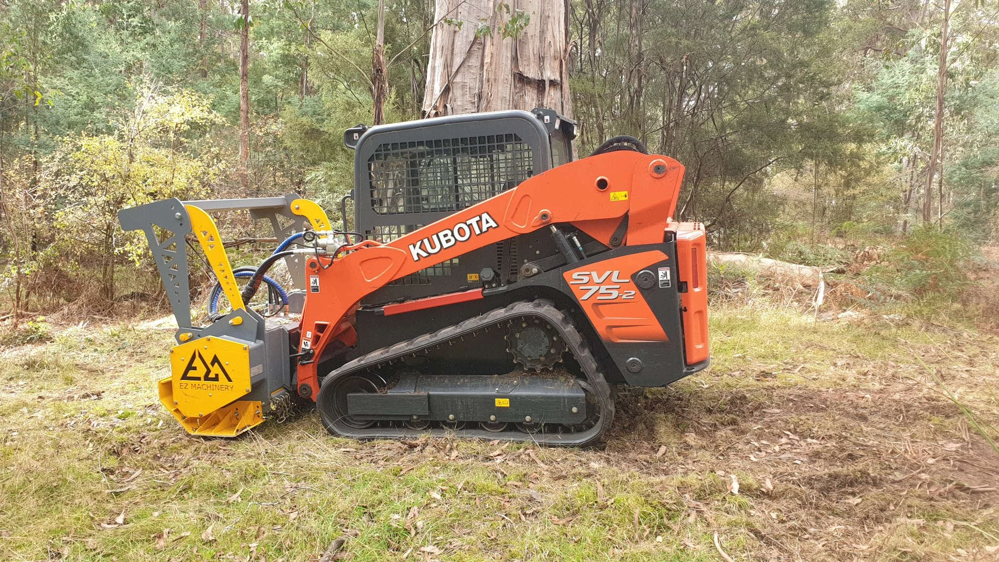 A Kubota SVL75-2 compact track loader is on a grassy, wooded outdoor area.