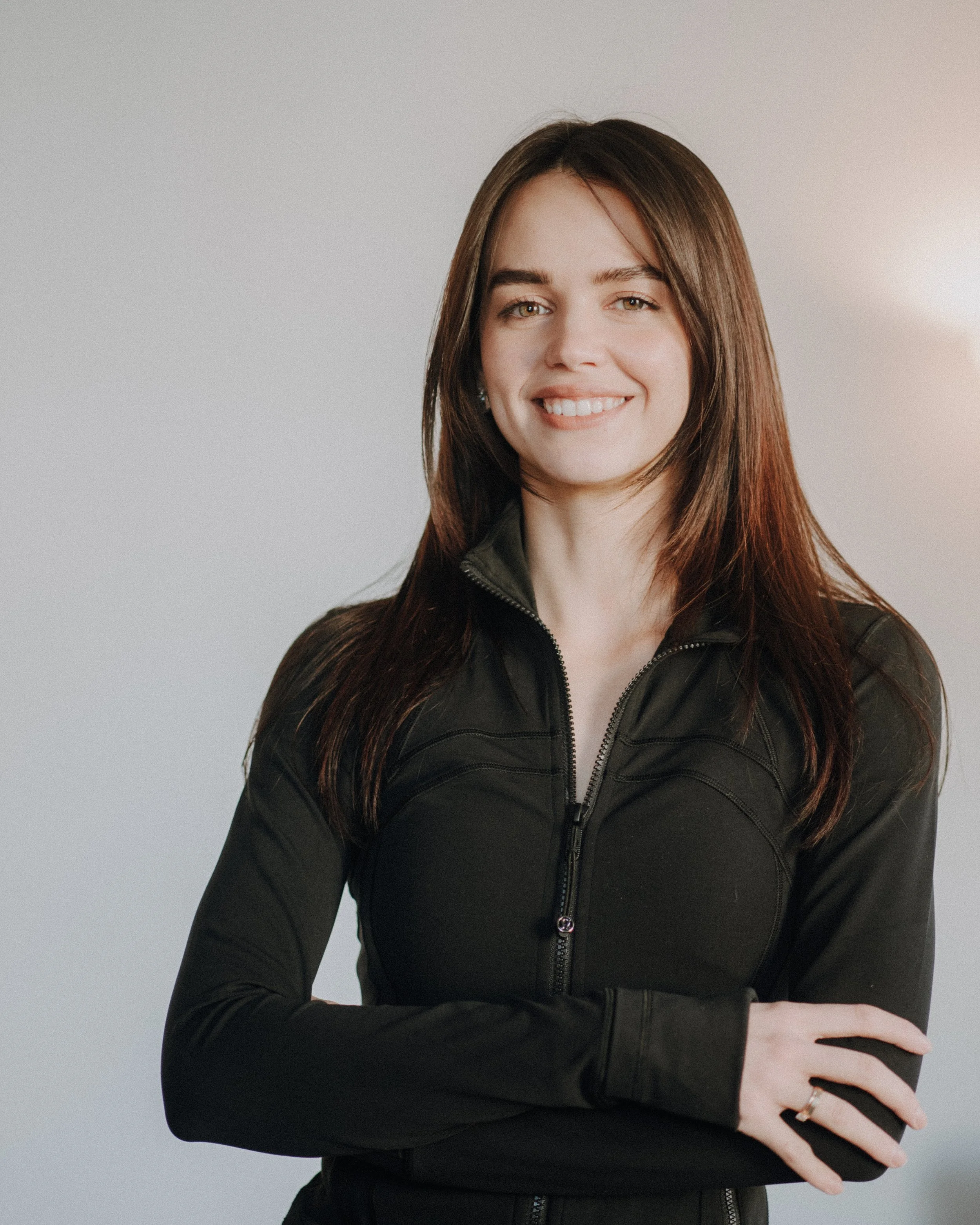 Unsere Trainerin Polina mit langen braunen Haaren, die ein weißes T-Shirt trägt, steht draußen vor einer Naturkulisse mit Bäumen und Pflanzen und lächelt.