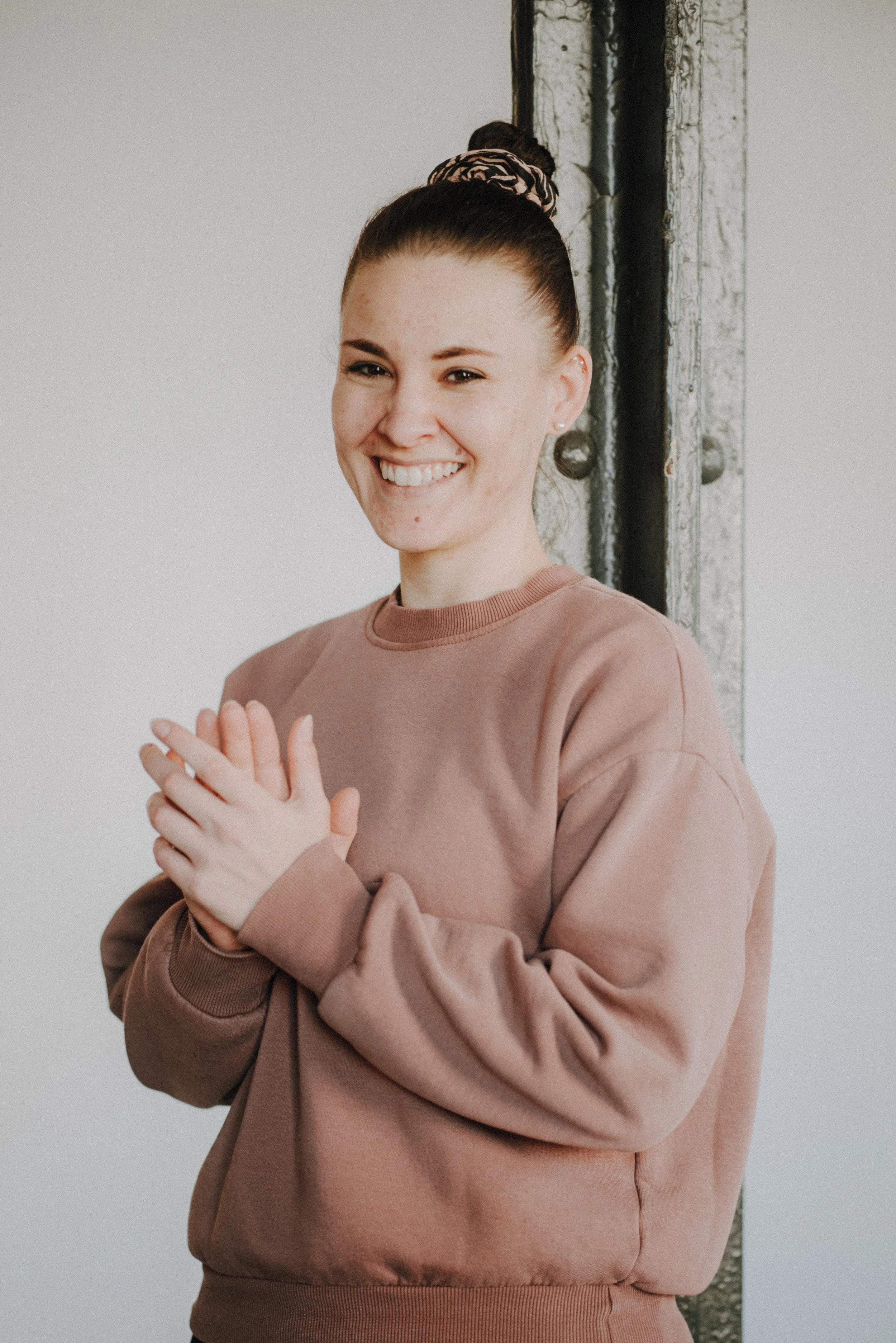Unsere Trainerin Julia mit dunklem Haar in einem Haarknoten, lächelnd. Trägt ein weißes T-Shirt, schwarze Shorts und steht im Freien vor einer Steinmauer.