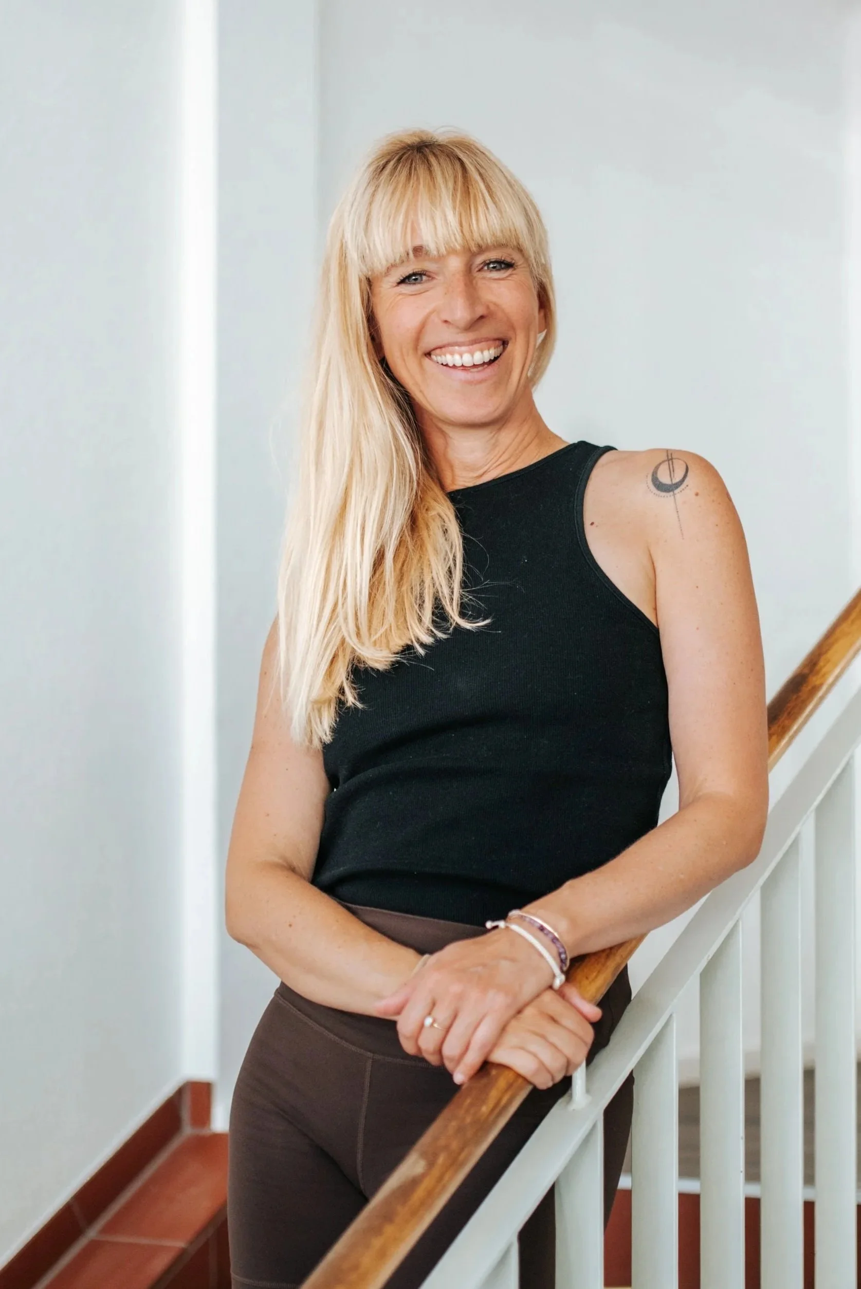 Owner & trainer Carla wears a white tank top and stands smiling in a bright room with her arms folded.