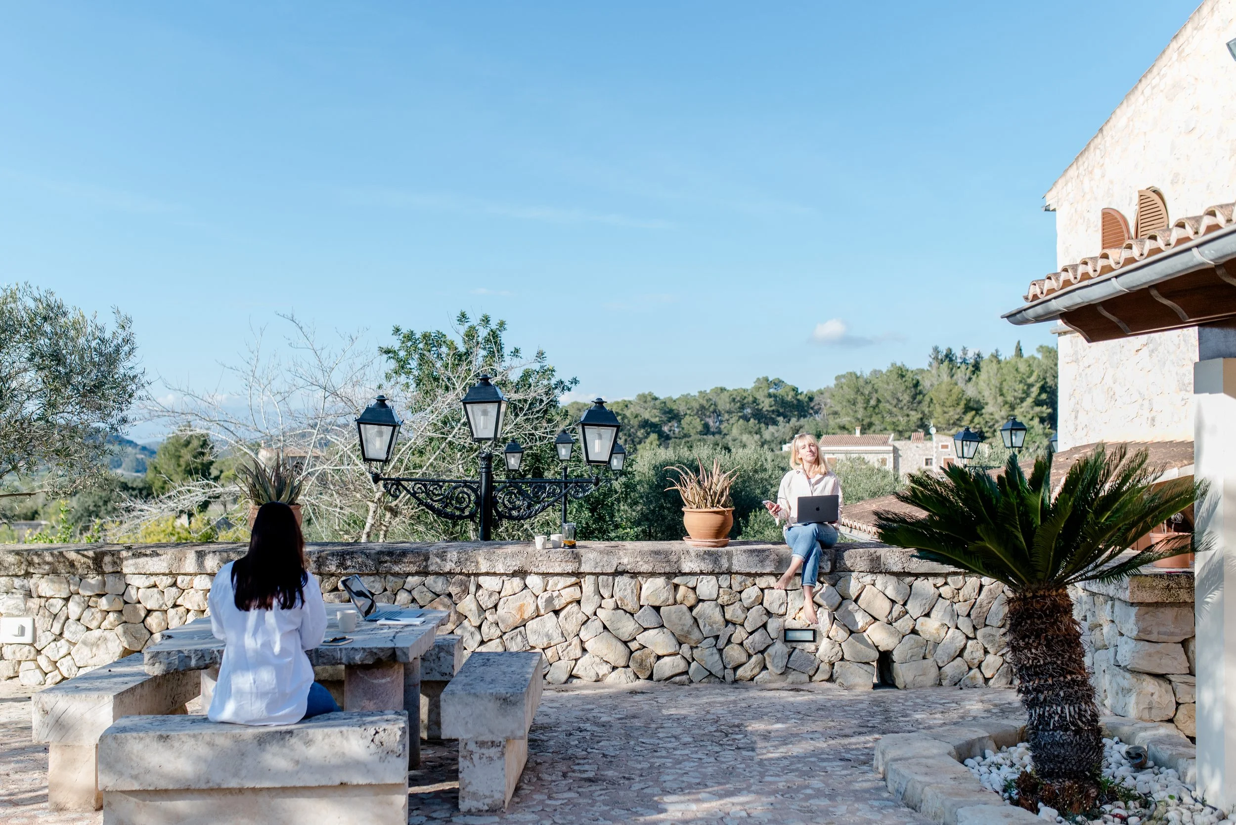 Carla and Anja are standing on a balcony next to an outdoor tree in yoga or fitness clothing, smiling at the camera.