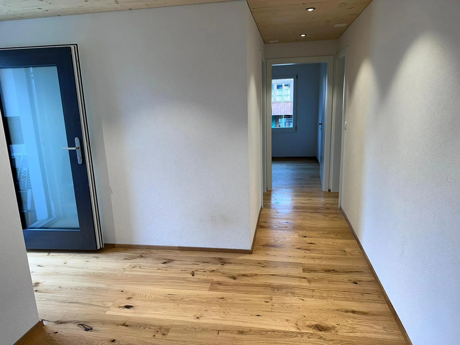 Ein heller Flur mit Holzboden, weißen Wänden und einem Fenster am Ende. Links befindet sich eine Tür mit Glas, rechts sind weitere Türen. Die Decke ist aus Holz.