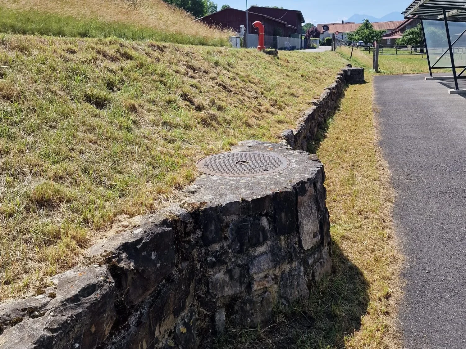 Ein Steinmauer mit einem Metallabdeckung, die einen Abfluss oder eine Kanalisation verbirgt, befindet sich neben einer asphaltierten Straße in einem ländlichen Gebiet mit Häusern und grünen Hügeln im Hintergrund.